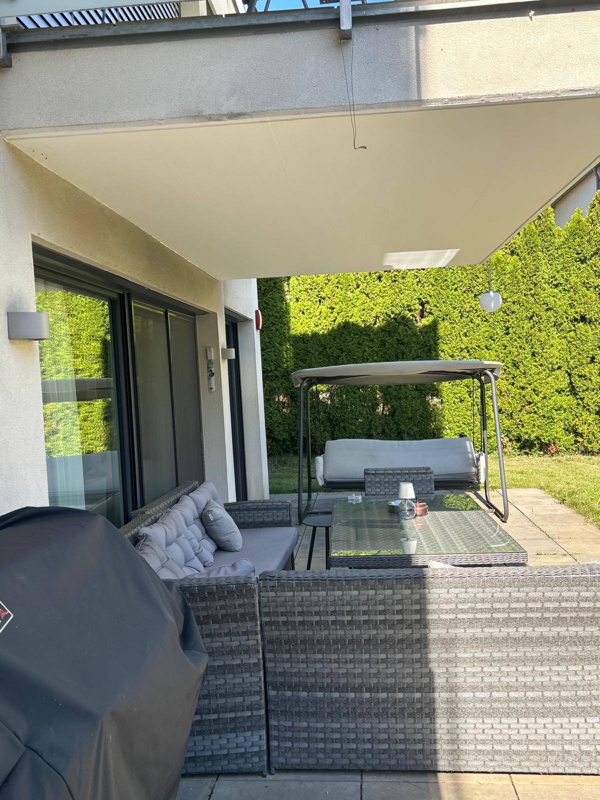 Sonnige Terrasse mit bequemen Gartenmöbeln, Schaukel und Blick auf die grüne Hecke.