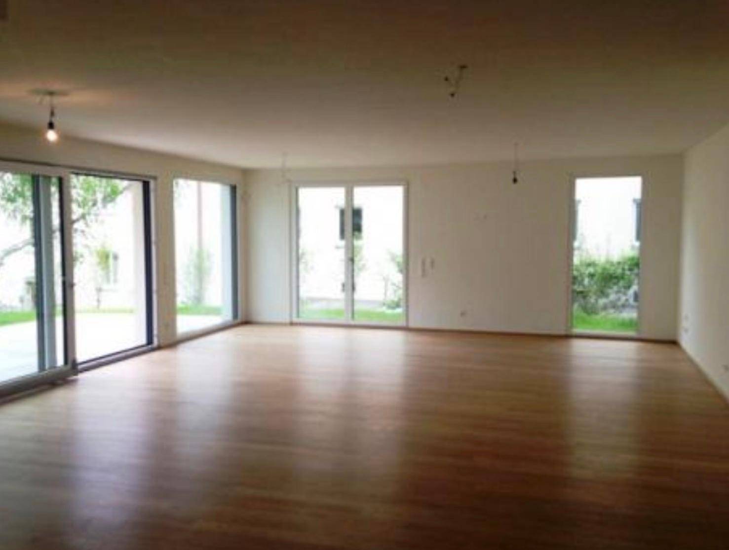 Heller, leerer Raum mit Holzboden und großen Fenstertüren zum Gartenbereich.