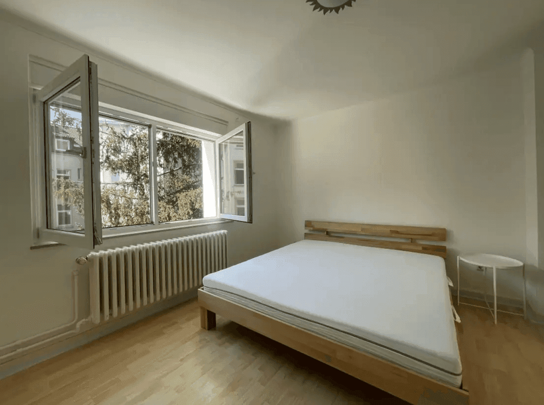 Schlafzimmer mit schlichtem Holzbettrahmen, Beistelltisch und großem Fenster mit Grünblick.