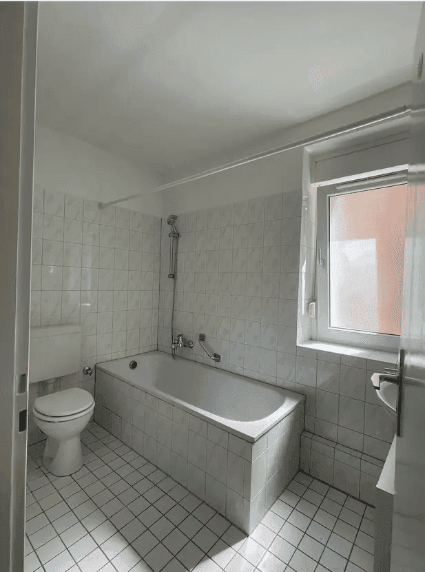 Helles Badezimmer mit Badewanne, WC und Fenster, komplett gefliest in Weiß.