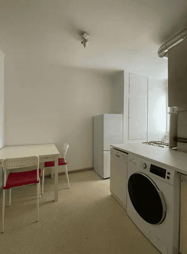 Funktionale Küche mit Kühlschrank, Waschmaschine und kleinem Essbereich am Fenster.