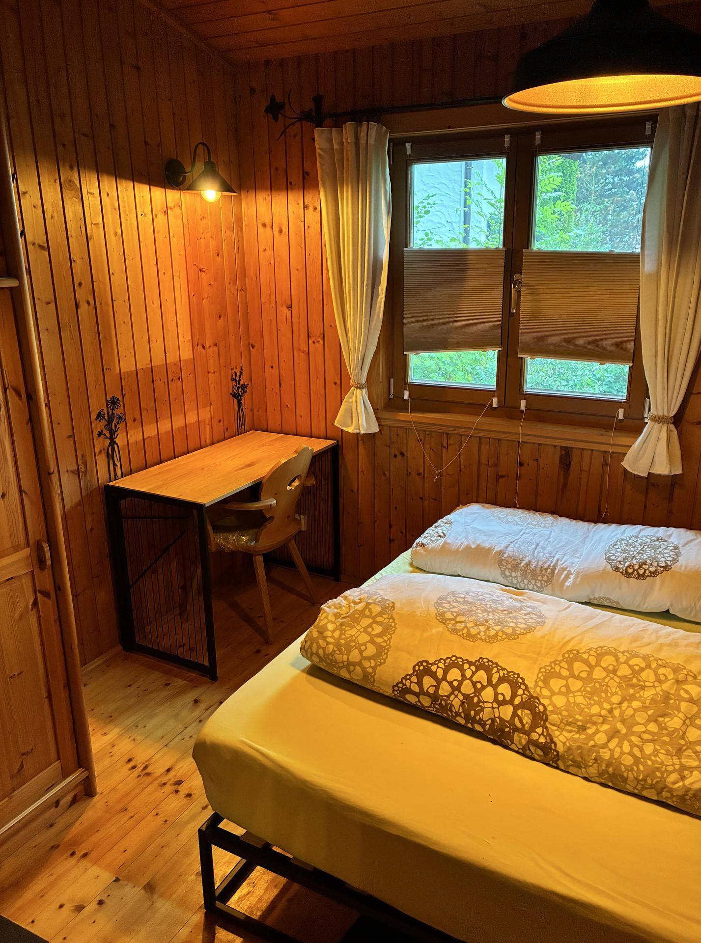 Schlichtes Schlafzimmer mit Holzwänden, einem Einzelbett und einem kleinen Schreibtisch am Fenster.
