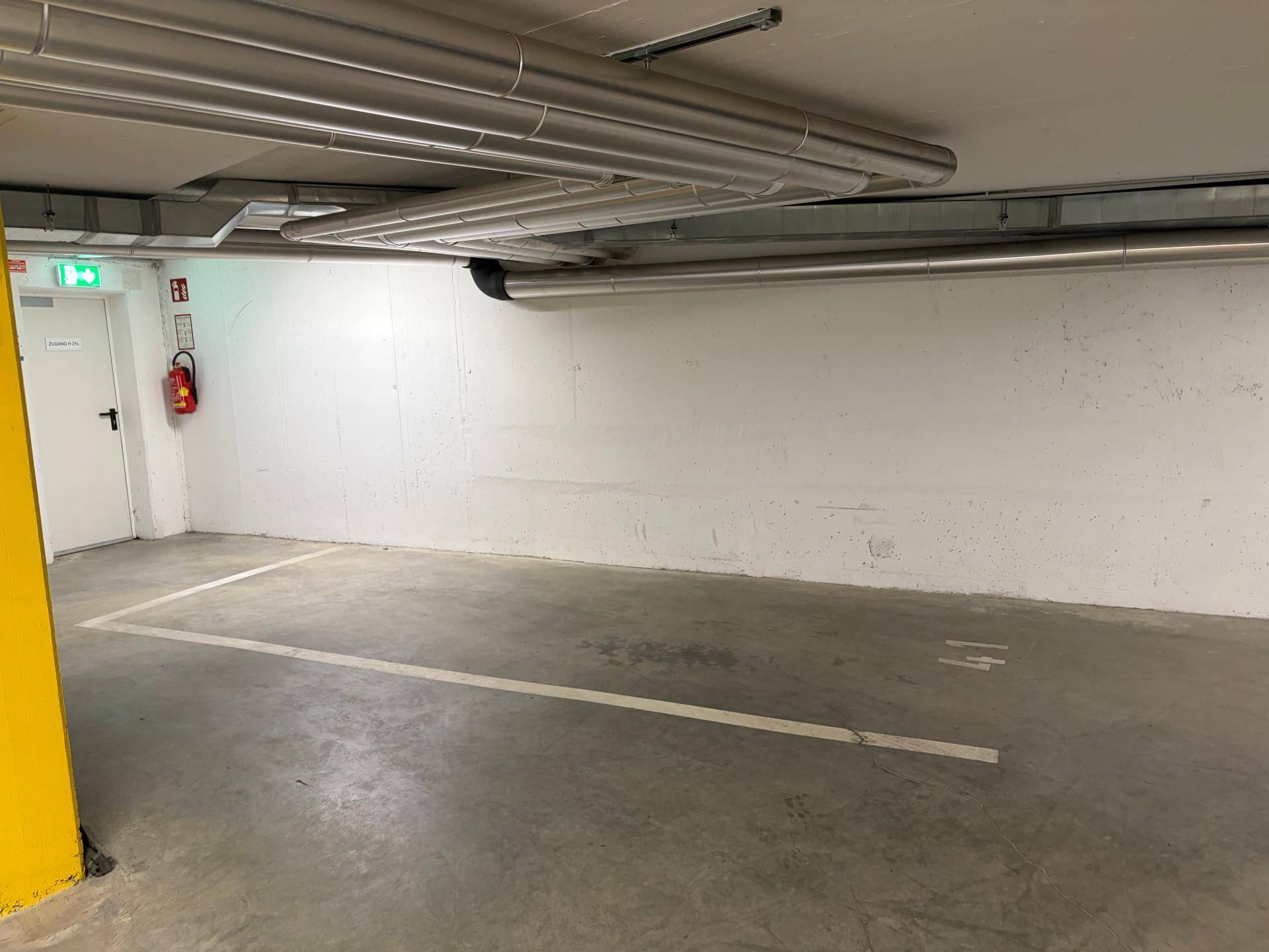 Geräumiger Tiefgaragenstellplatz mit Betonboden, einem gelben Pfeiler und sichtbaren Deckenleitungen, ideal für sicheres Parken.