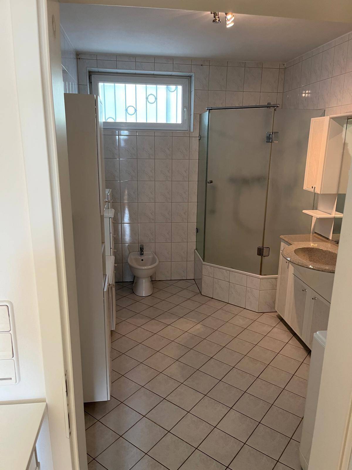 Helles Badezimmer mit Duschkabine, Bidet, Fenster und weißen Fliesen.