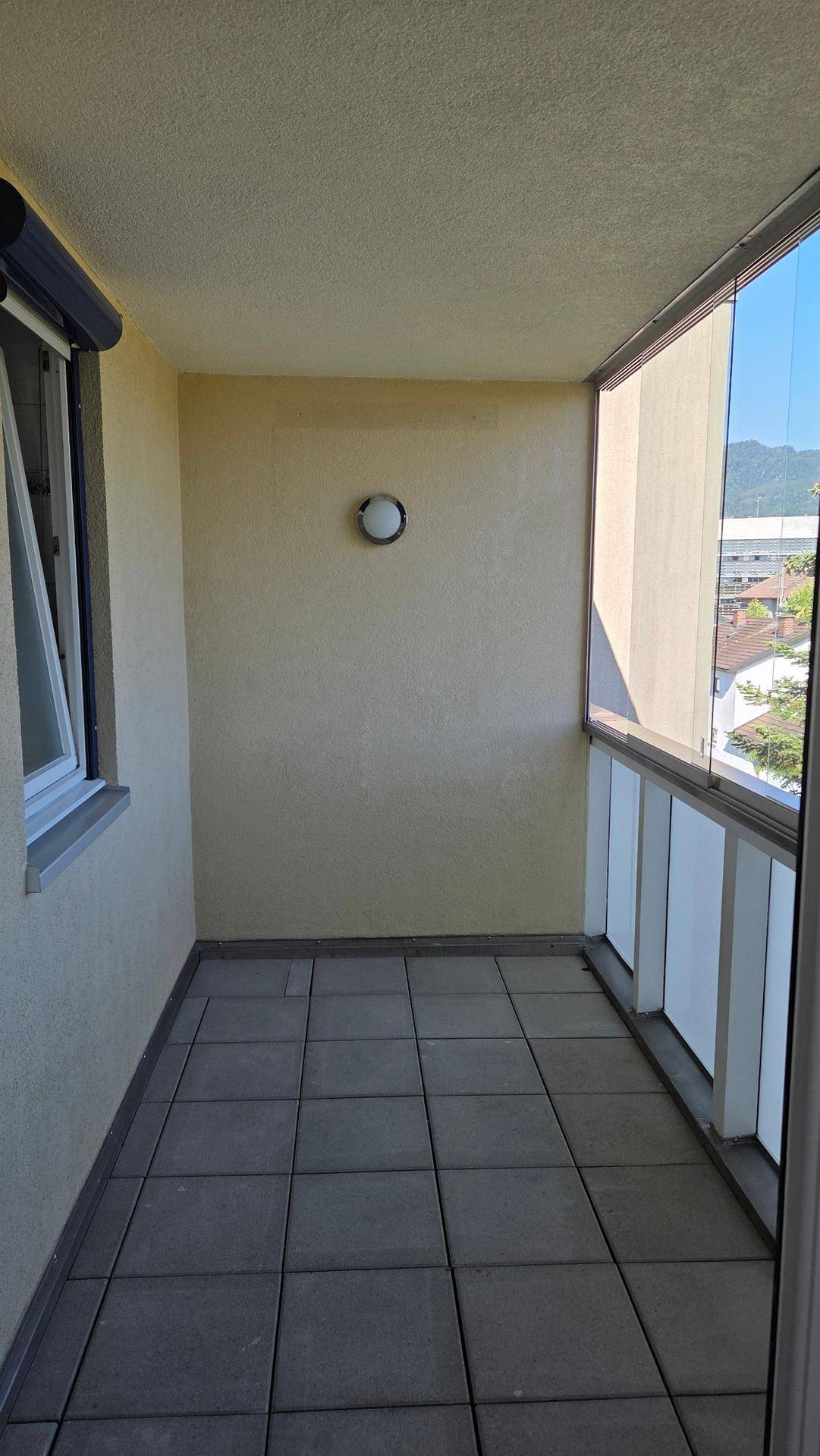 Kleiner, gefliester Balkon mit Glasbrüstung und Rollladen am Fenster.