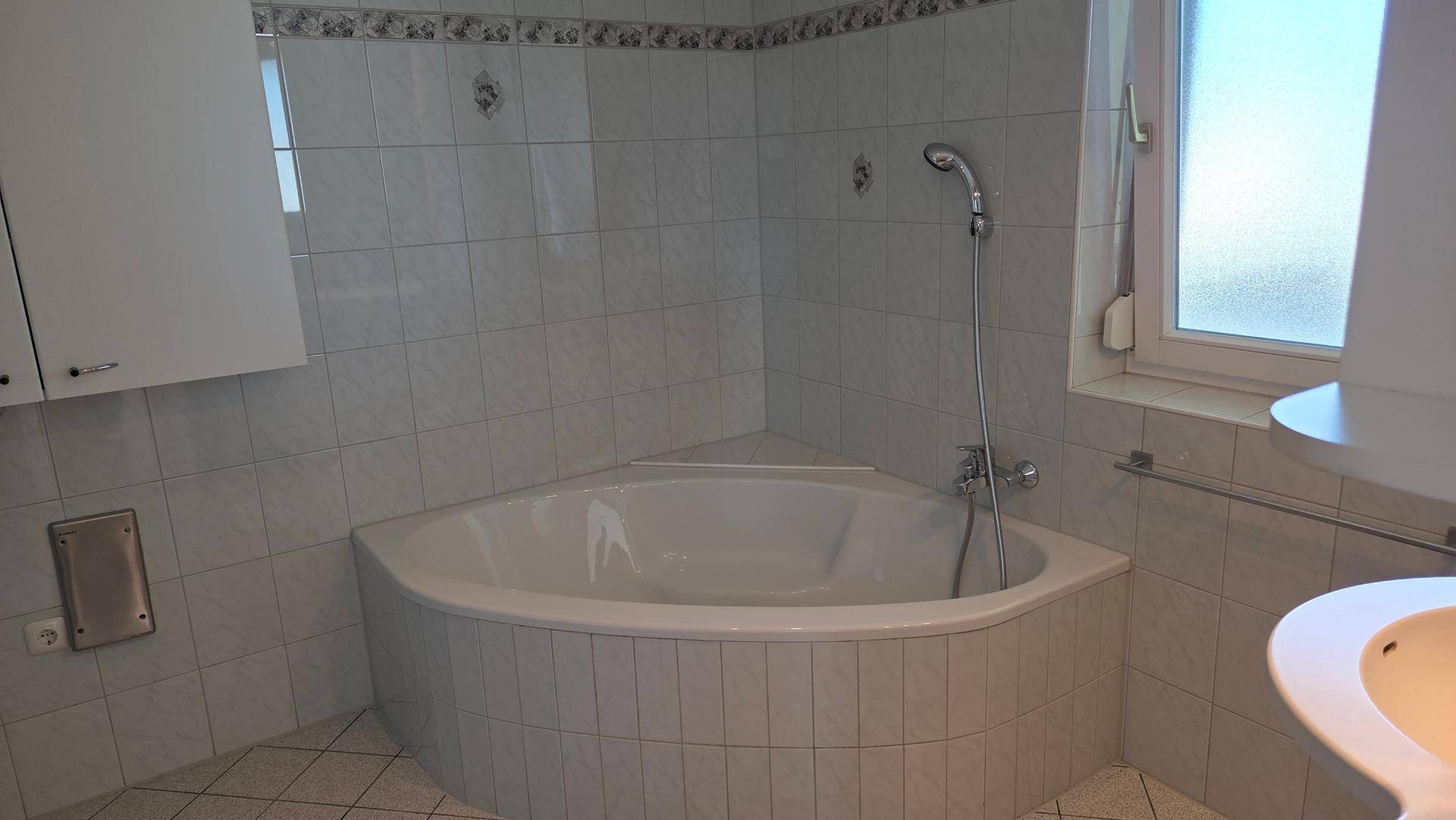 Badezimmer mit Eckbadewanne, Fliesenboden und Fenster für natürliche Belüftung.