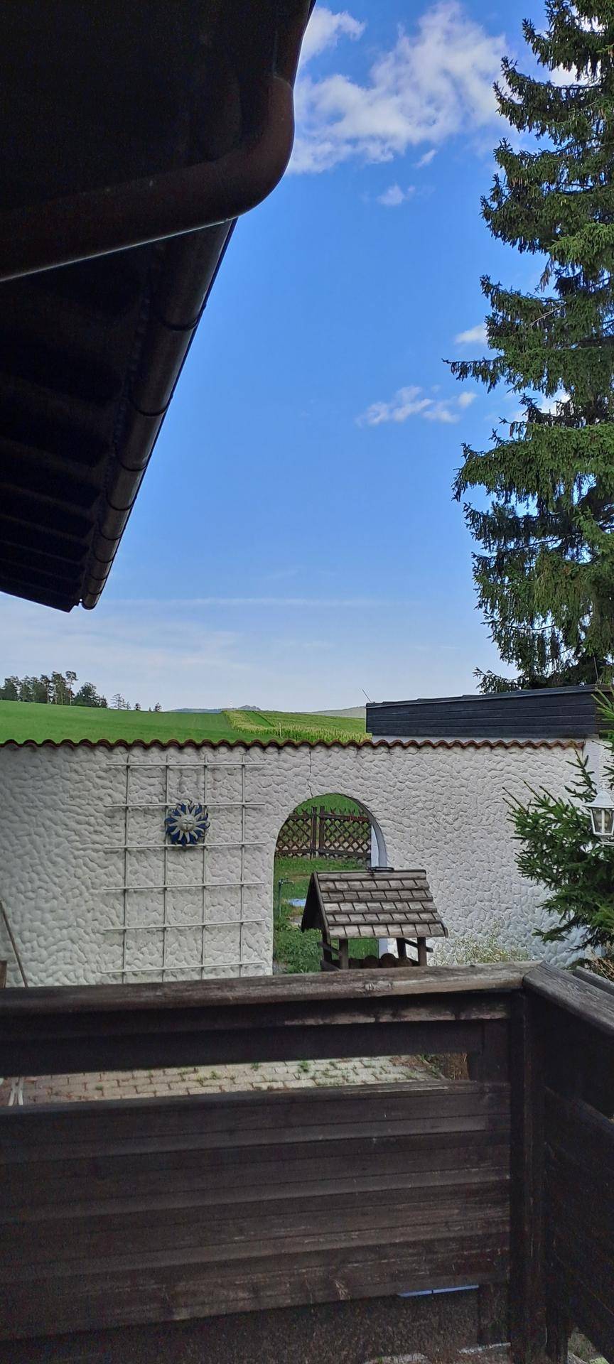 Blick über eine weiße Mauer mit Torbogen auf grüne Felder und den blauen Himmel.