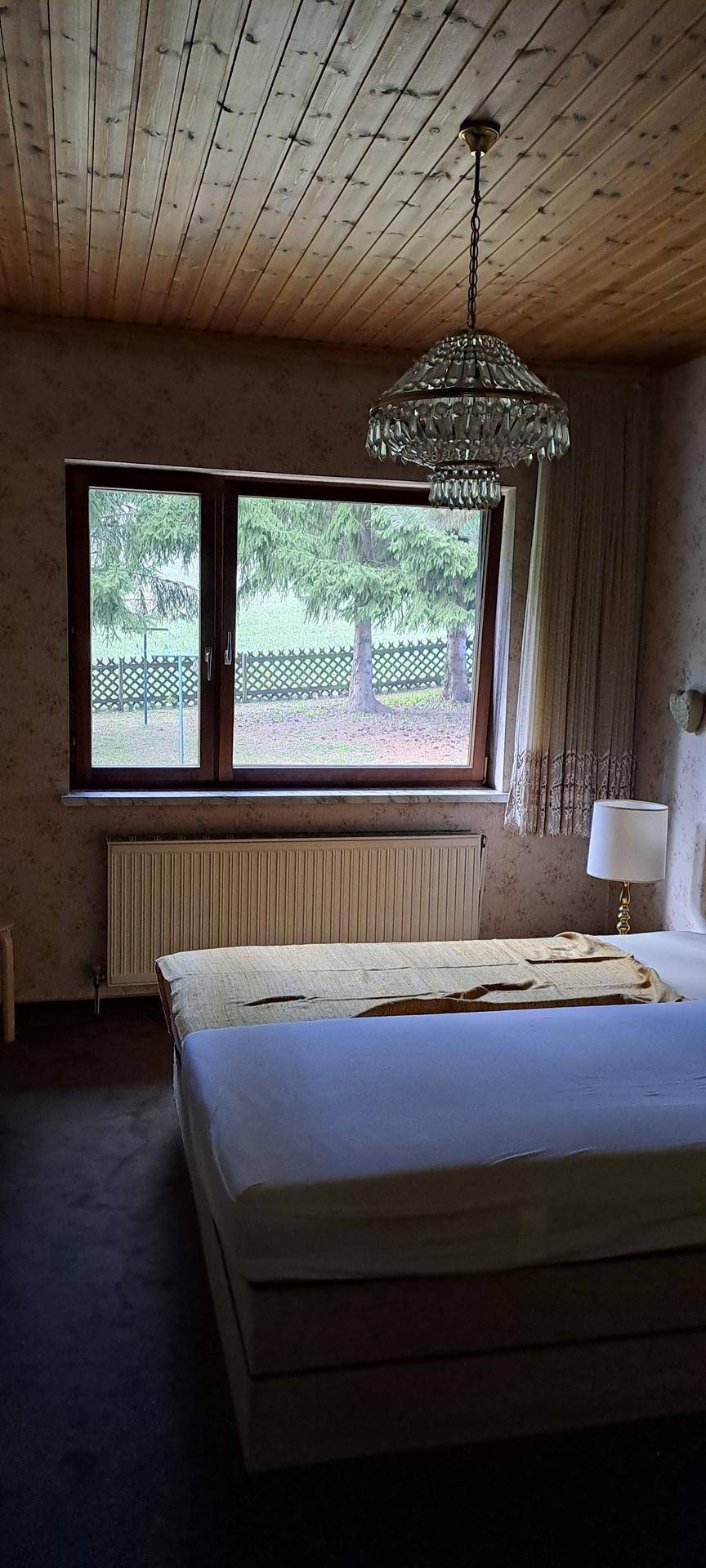 Schlafzimmer mit Doppelbett, Holzdecke, Fenster mit Blick ins Grüne und Kronleuchter.