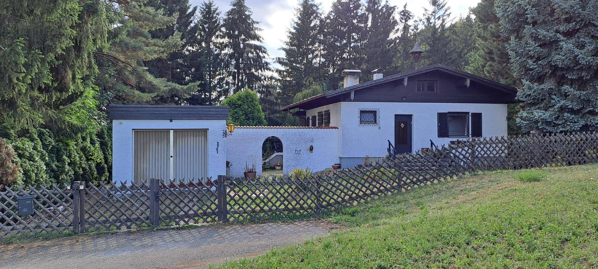 Freistehendes weißes Haus mit Garage und Holzzaun, eingebettet in eine grüne Landschaft.