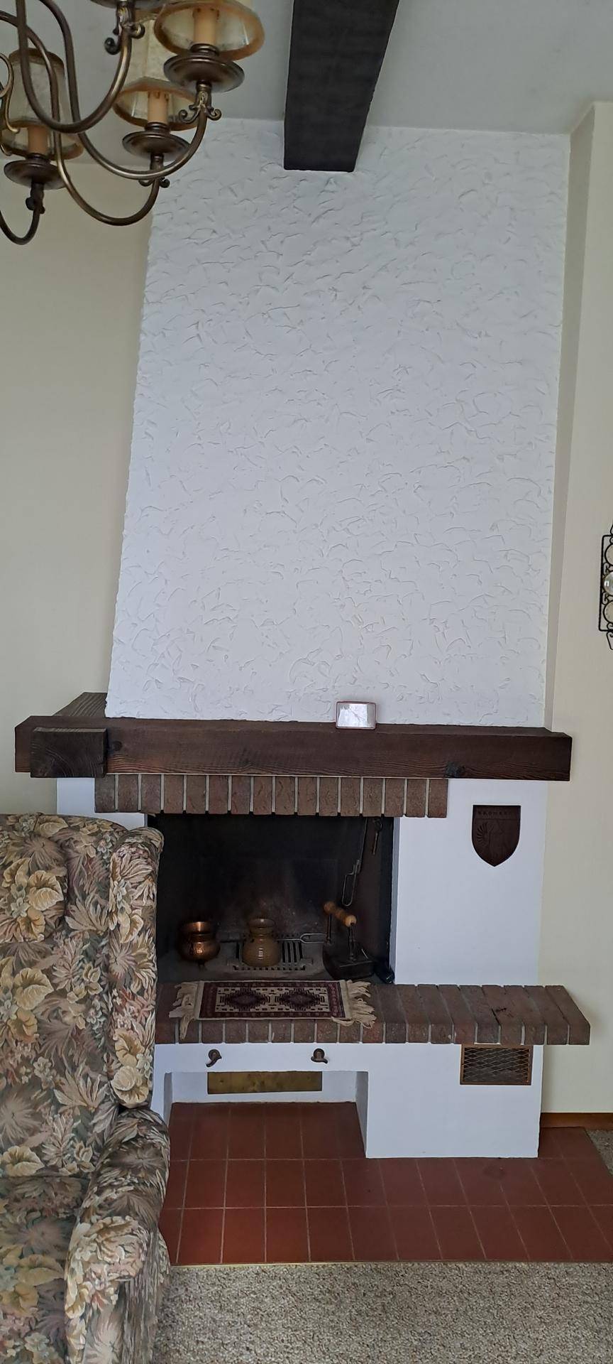 Traditioneller Kamin mit weißer Strukturputzoberfläche und Holzbalken, neben einem Sessel.