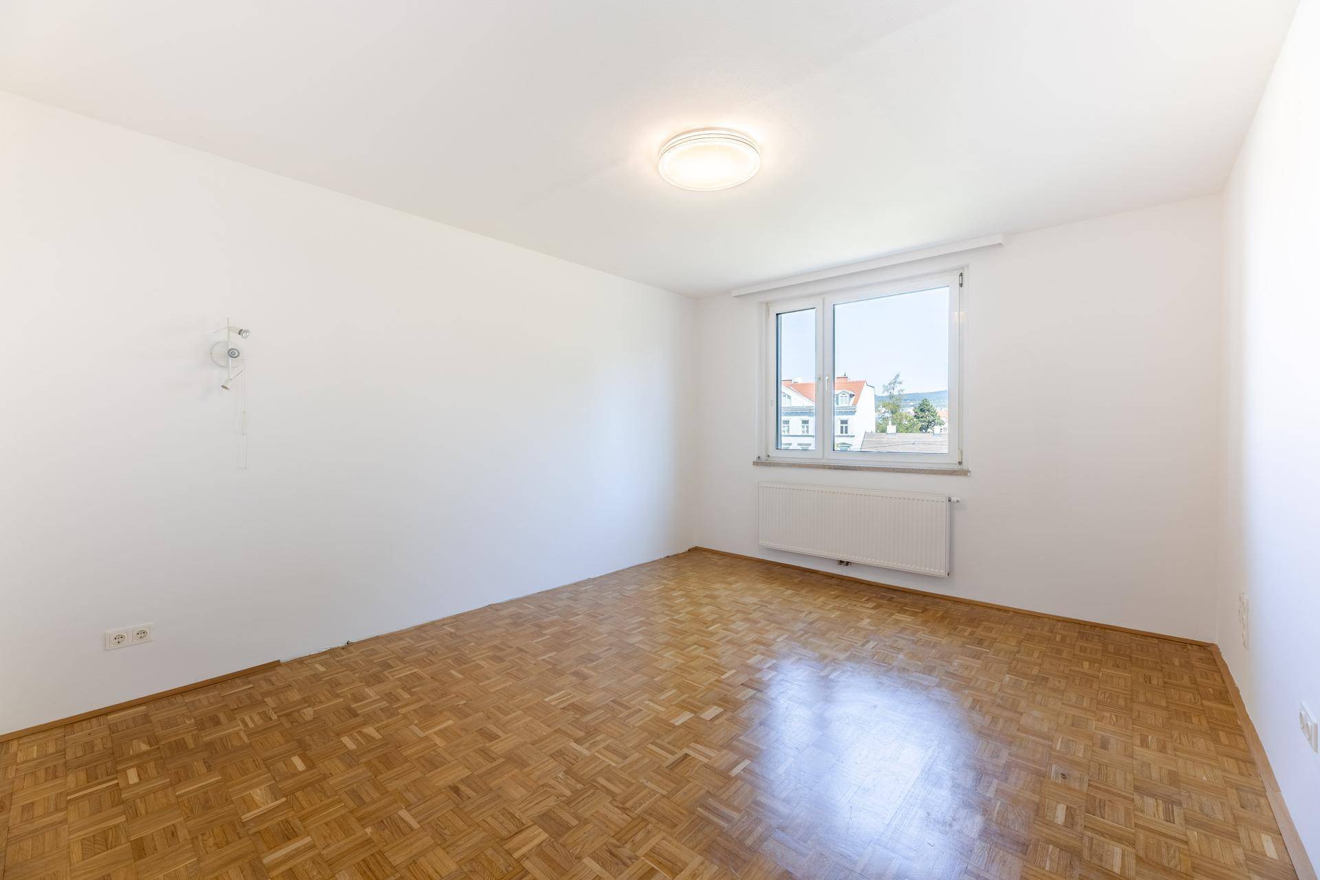 Helles Schlafzimmer mit Parkettboden, weiß gestrichenen Wänden und Blick auf die Umgebung.