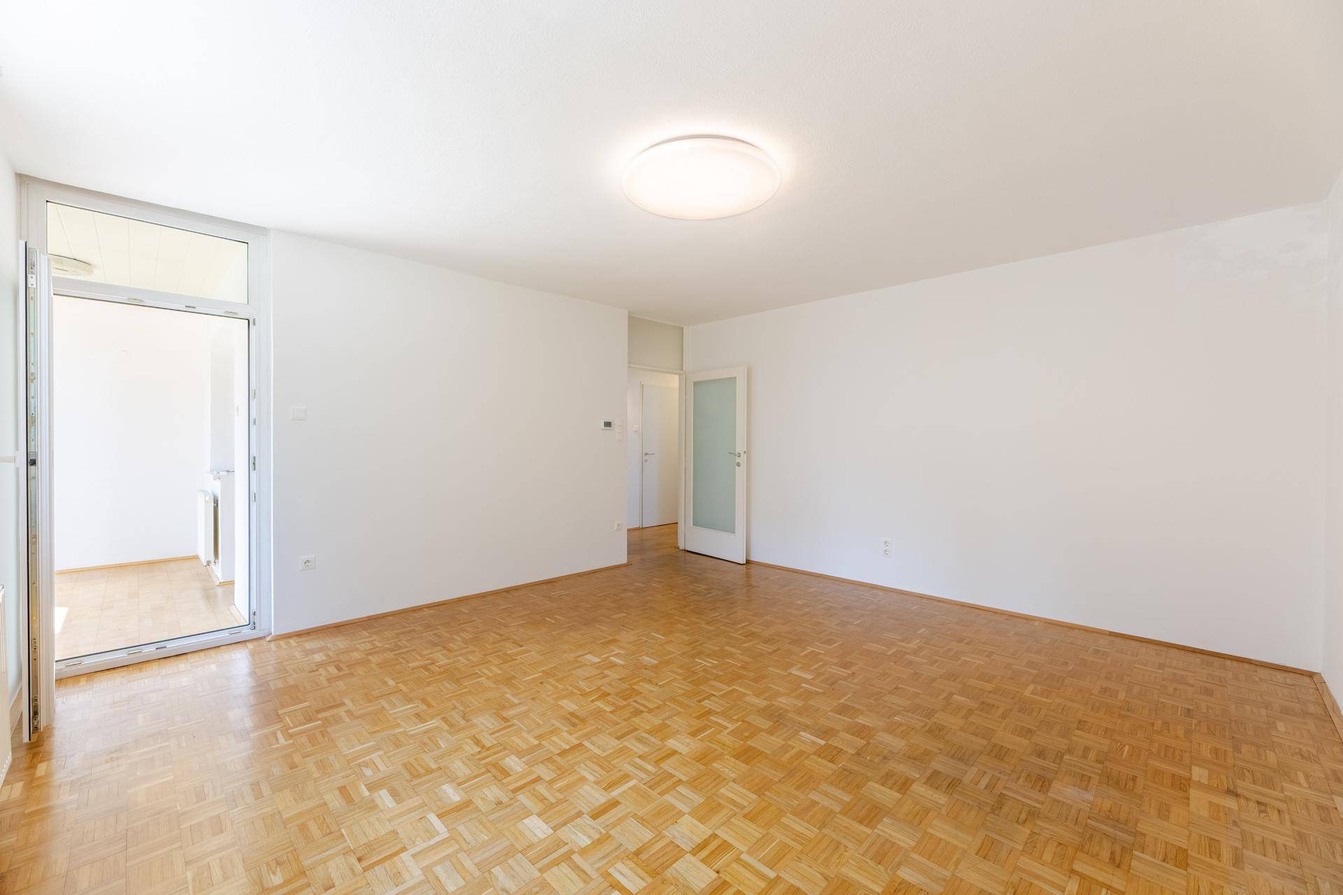 Geräumiges, leeres Wohnzimmer mit hellem Parkettboden und Zugang zu einem Außenbereich.