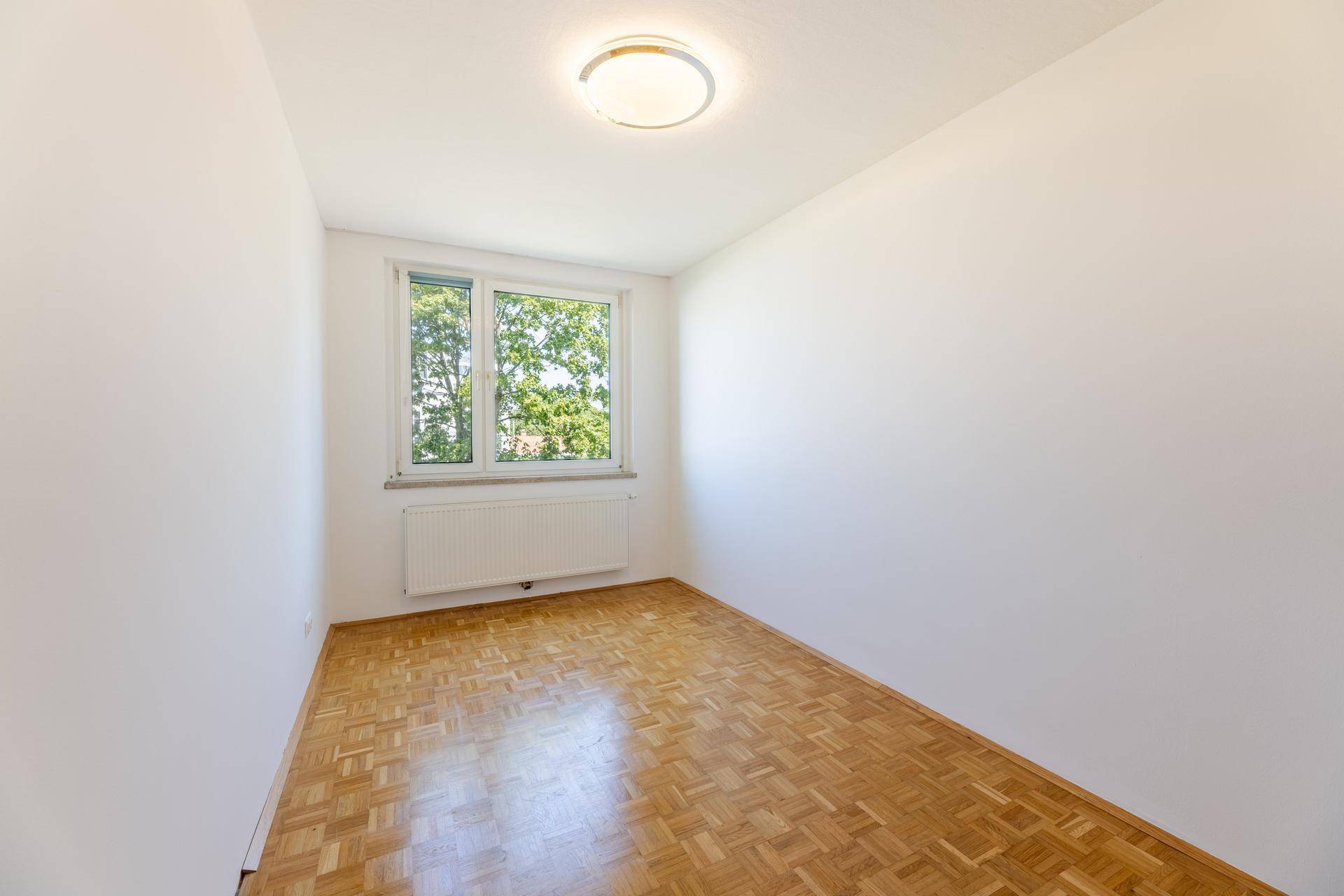 Kleineres Zimmer mit Parkettboden, weißen Wänden und Fenster mit Blick ins Grüne.