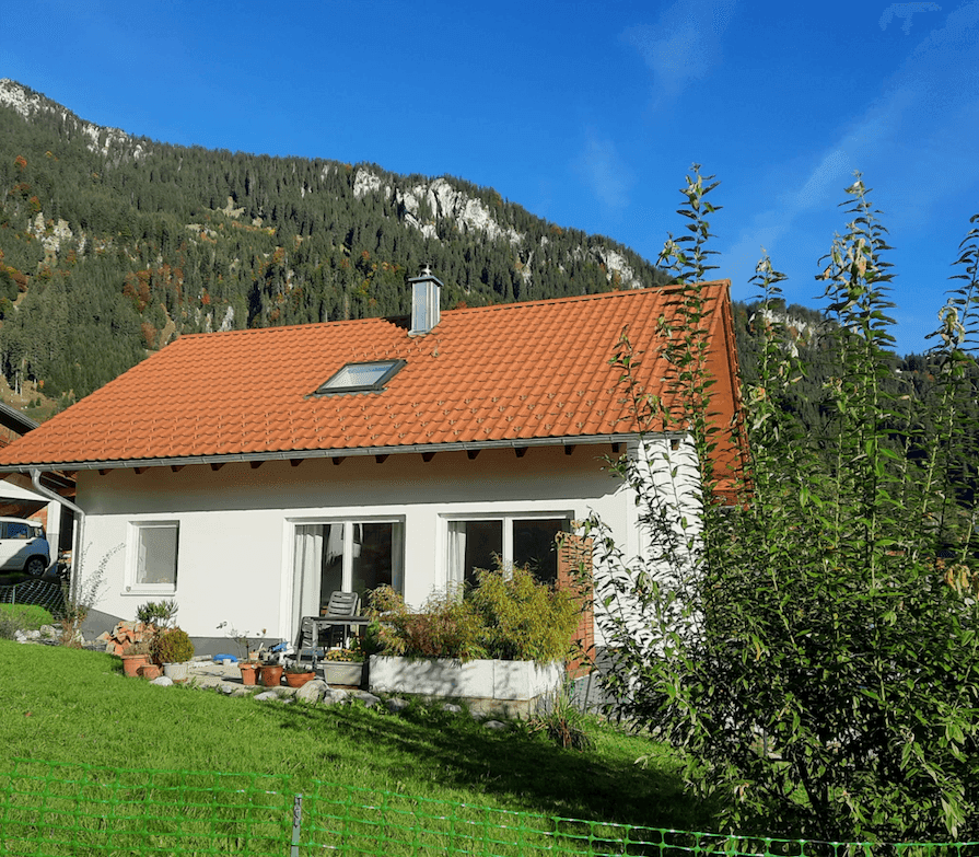 Hausrückseite mit kleiner Terrasse und Gartenbereich, umgeben von grüner Natur und Bergen.