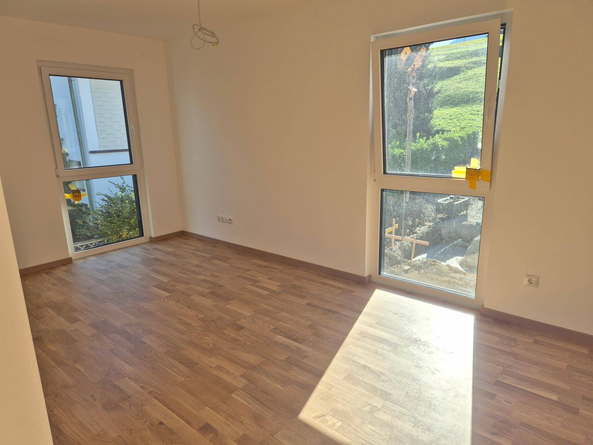 Helles Schlafzimmer mit Holzboden und zwei großen Fenstern, die einen Blick ins Grüne bieten.