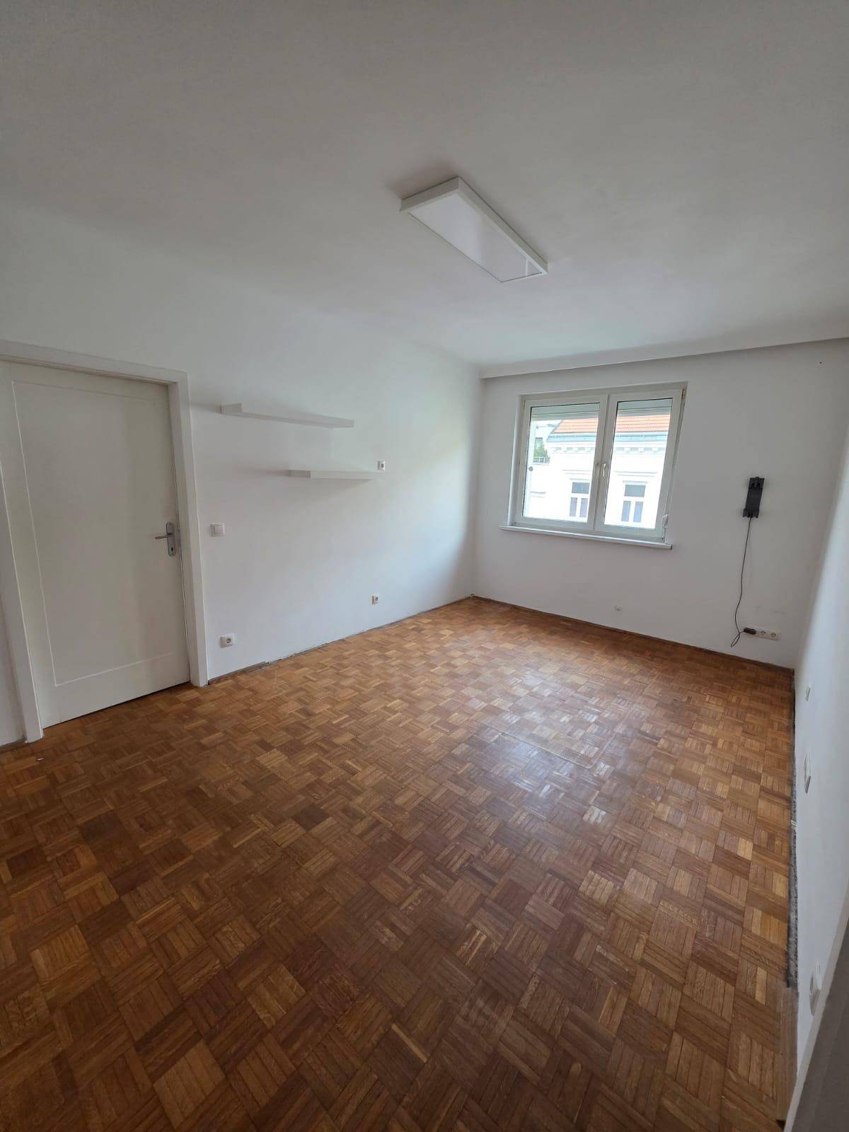 Geräumiger, heller Raum mit klassischem Parkettboden und einem Fenster.