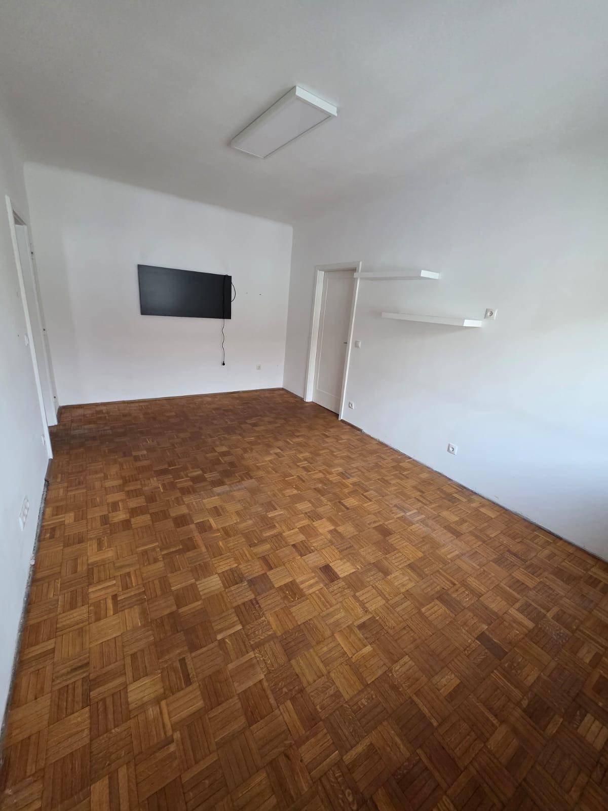 Heller Wohnraum mit Parkettboden, Wandregalen und montiertem Fernseher.