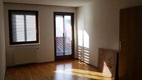 Leerer Raum mit Parkettboden, Fenster mit Jalousien und Zugang zu einem sonnigen Balkon.