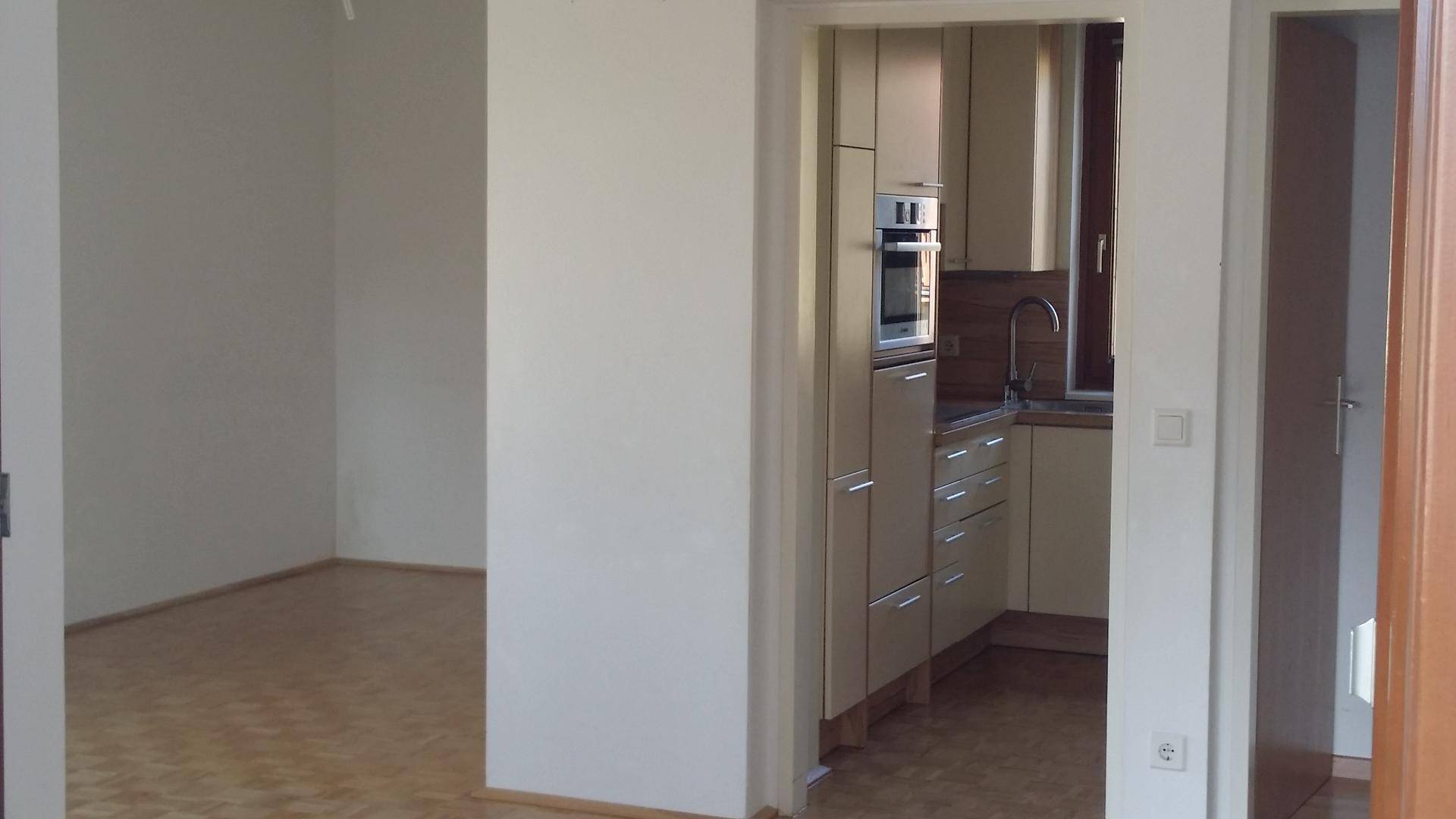 Blick von einem leeren Raum in eine helle Küche mit modernen Einbauschränken und Geräten.