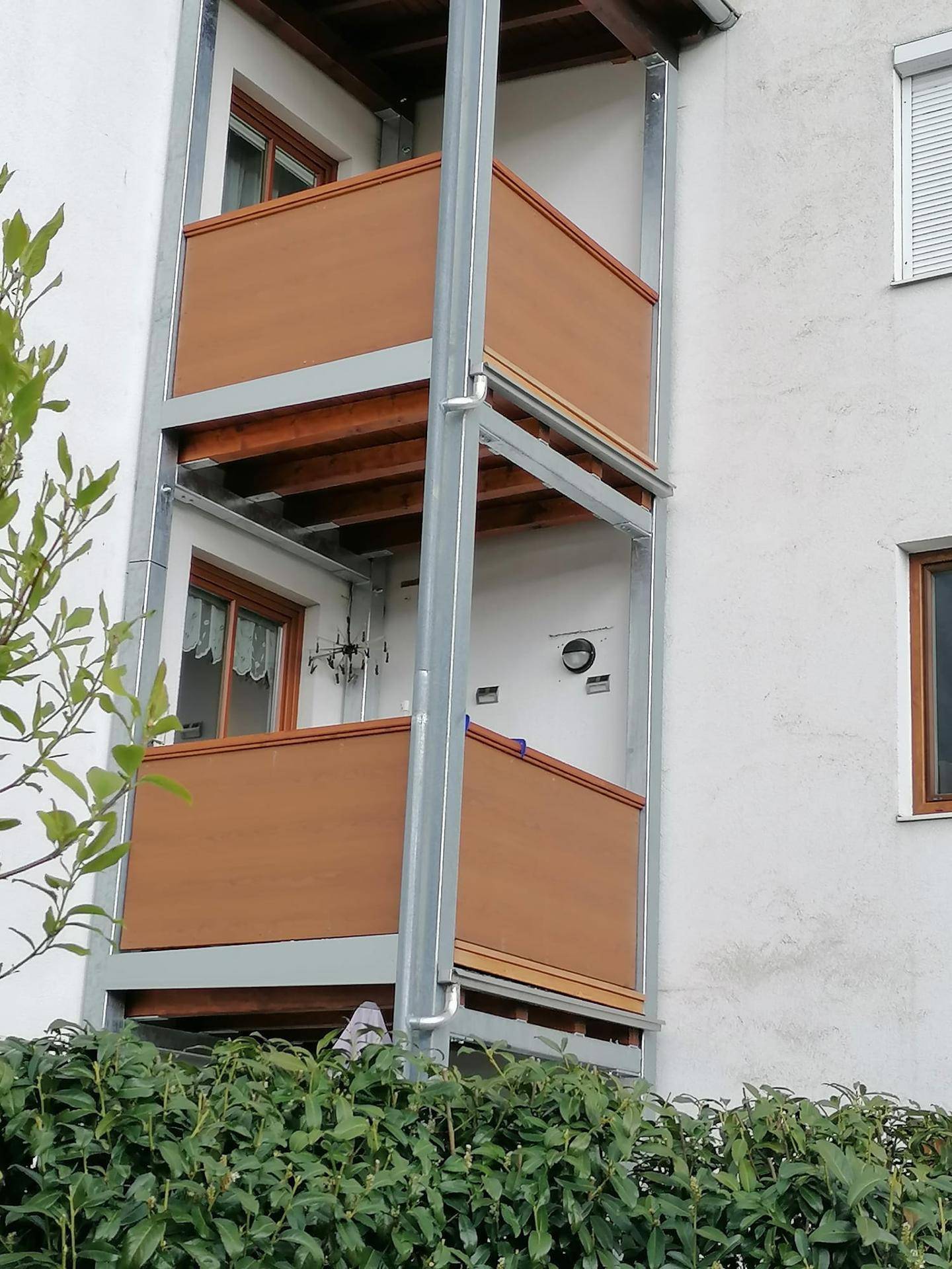 Außenansicht eines Gebäudes mit zwei übereinanderliegenden Balkonen und grüner Hecke im Vordergrund.