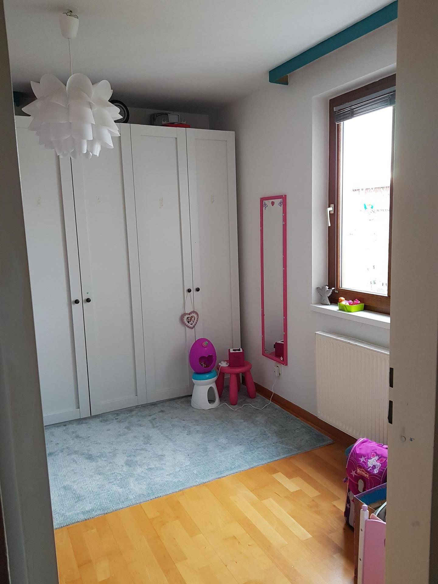 Helles Kinderzimmer mit großem weißen Kleiderschrank, Teppich und einem Fenster.