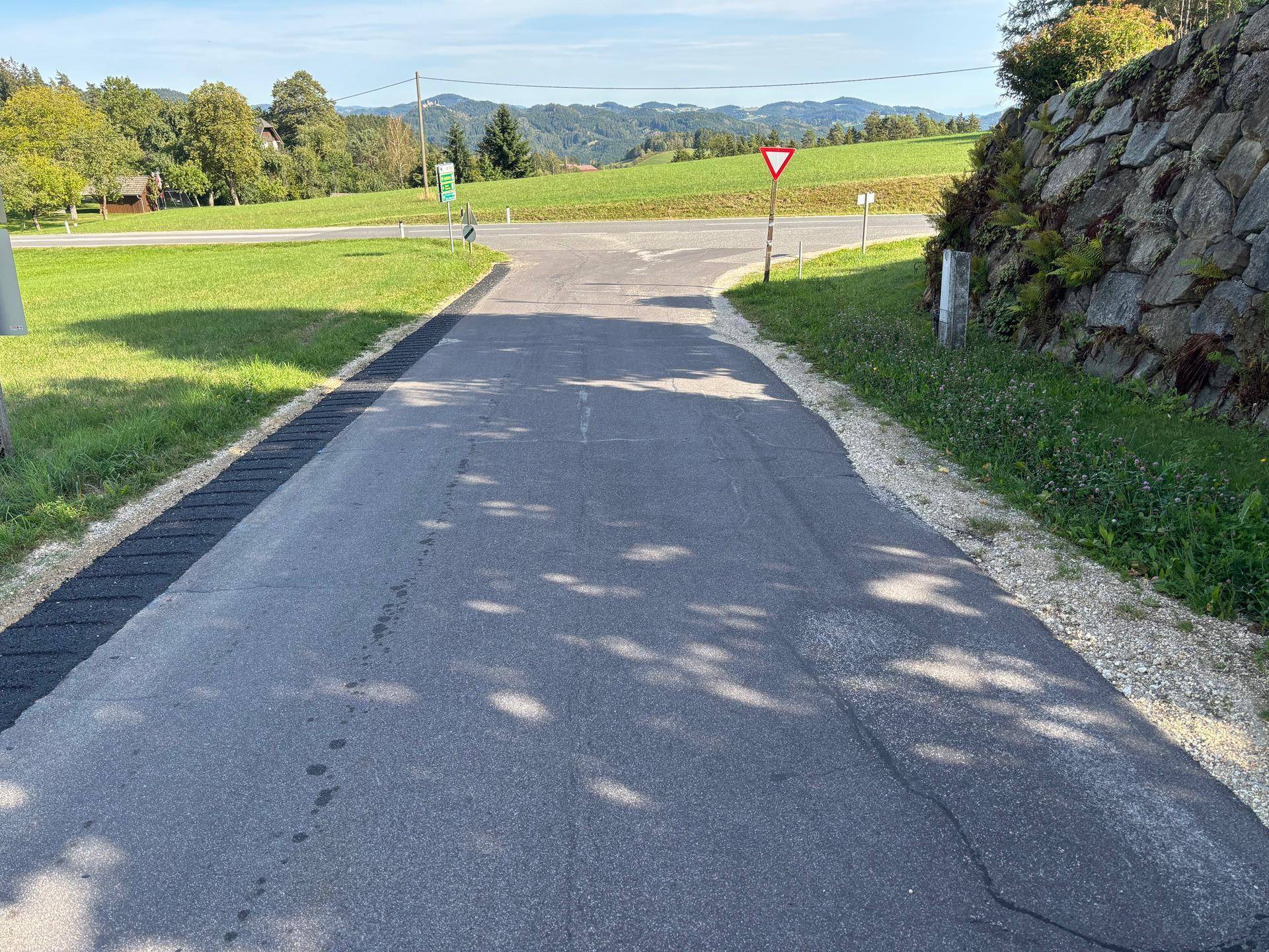 Asphaltierte Straße mit einer Kreuzung und einem Vorfahrt-achten-Schild, umgeben von grünen Hügeln und einer Steinmauer.
