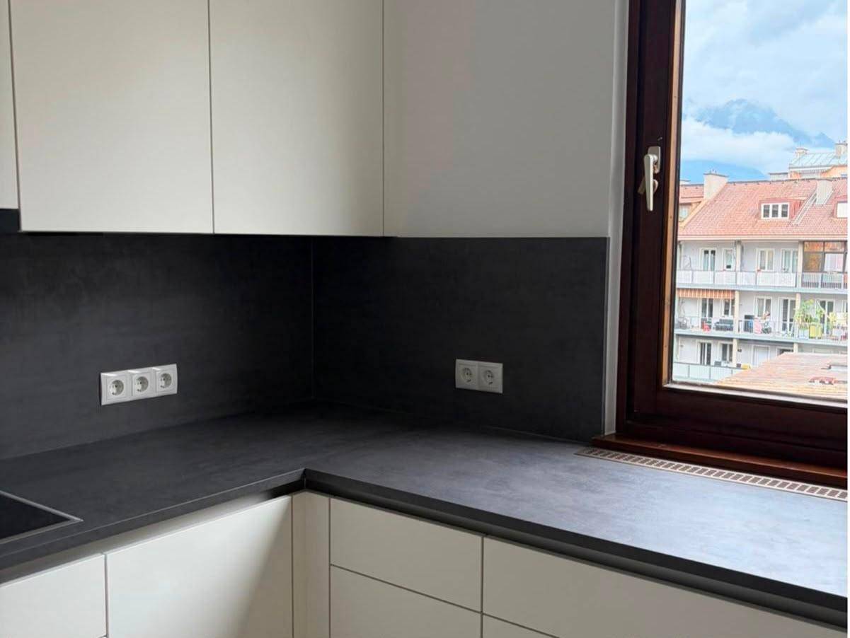 Moderne Küche mit dunkler Arbeitsplatte, weißen Schränken und einem Fenster mit Blick auf die Dächer.