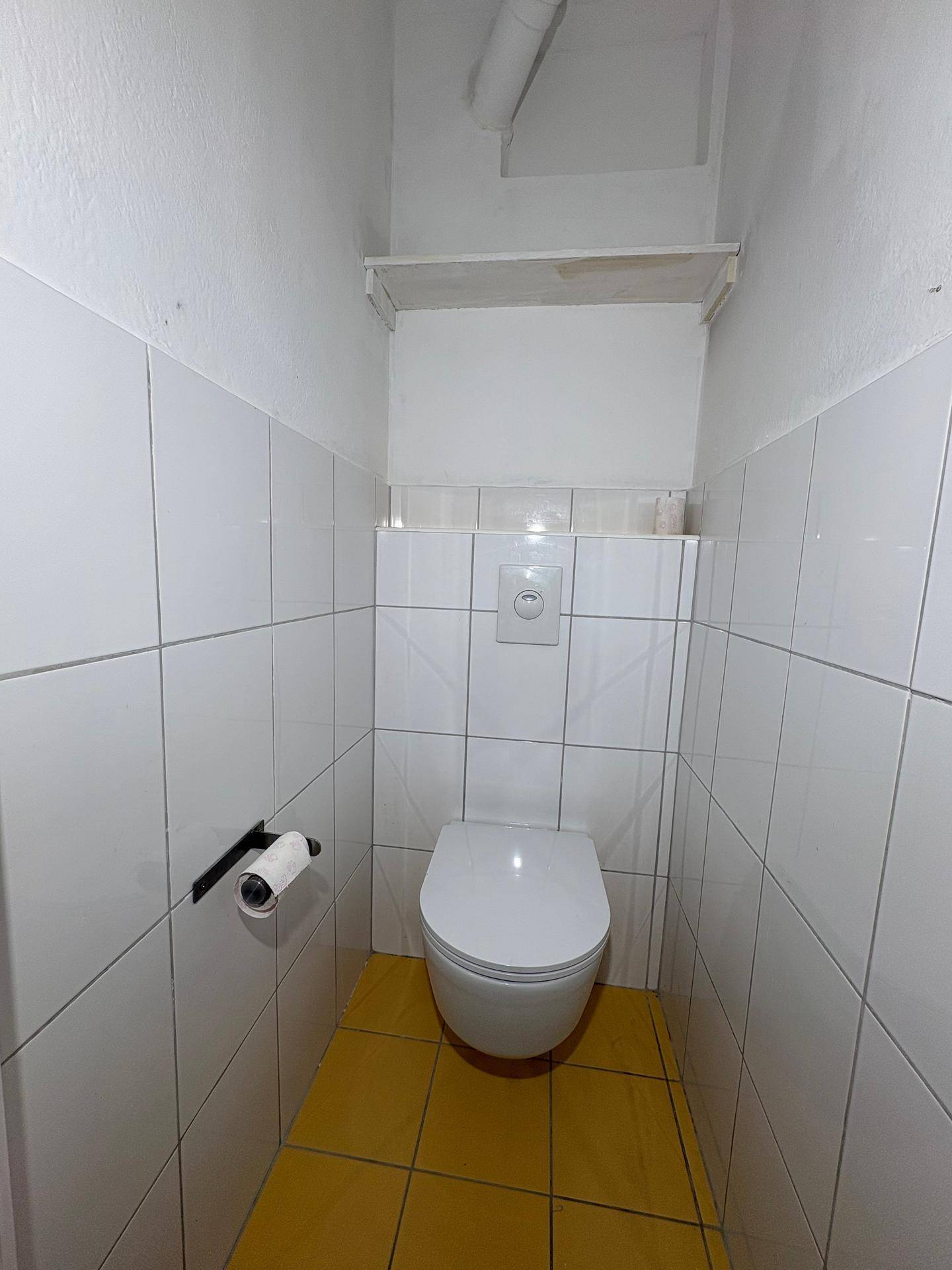 Kleines Gäste-WC mit weißen Wandfliesen, gelbem Boden und wandhängendem WC.
