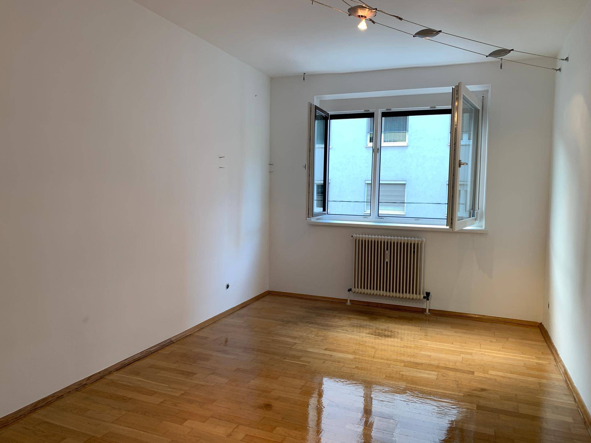 Leeres Zimmer mit hellem Parkettboden, weißen Wänden und einem großen Fenster, ideal als Schlafzimmer.