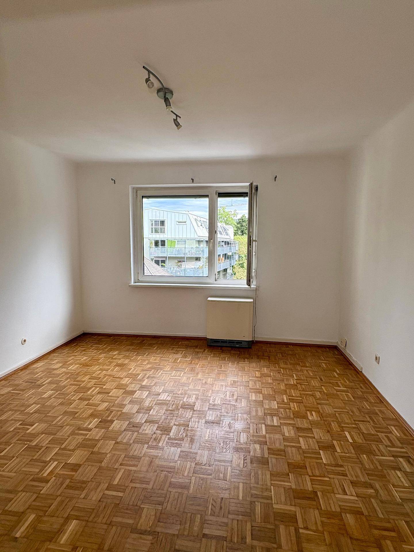 Geräumiges, leeres Zimmer mit hellem Parkettboden und einem großen Fenster, das viel Tageslicht hereinlässt.