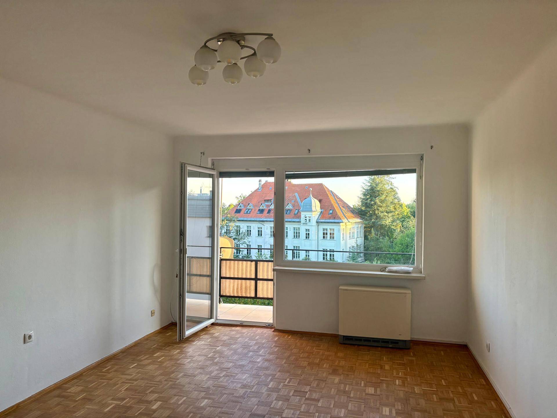 Helles, leeres Wohnzimmer mit Parkettboden, Zugang zum Balkon und Blick auf ein historisches Gebäude.