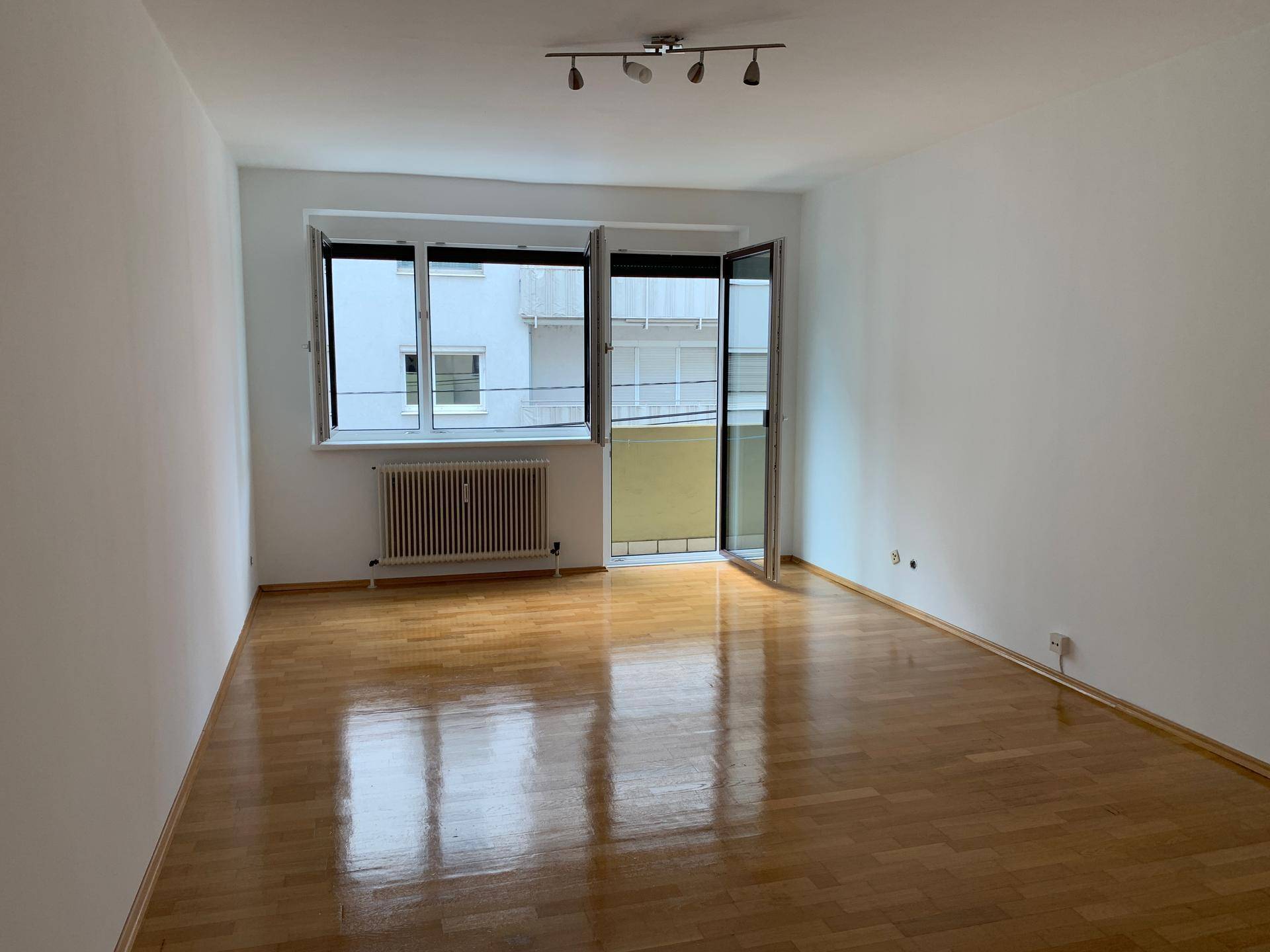 Helles, leeres Wohnzimmer mit glänzendem Parkettboden, großem Fenster und Zugang zum Balkon.