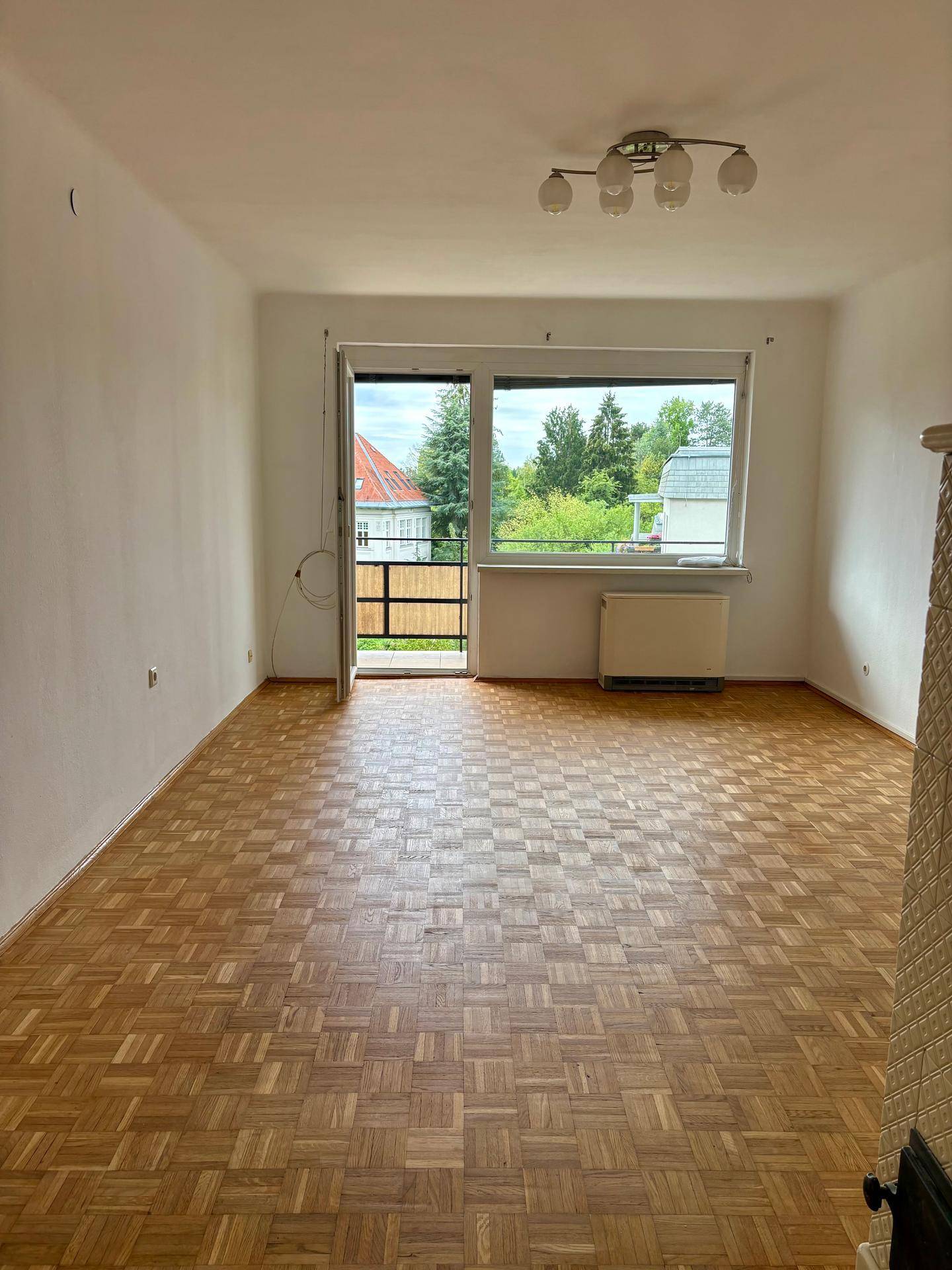 Helles, leeres Wohnzimmer mit Parkettboden, großem Fenster und Zugang zu einem kleinen Balkon.