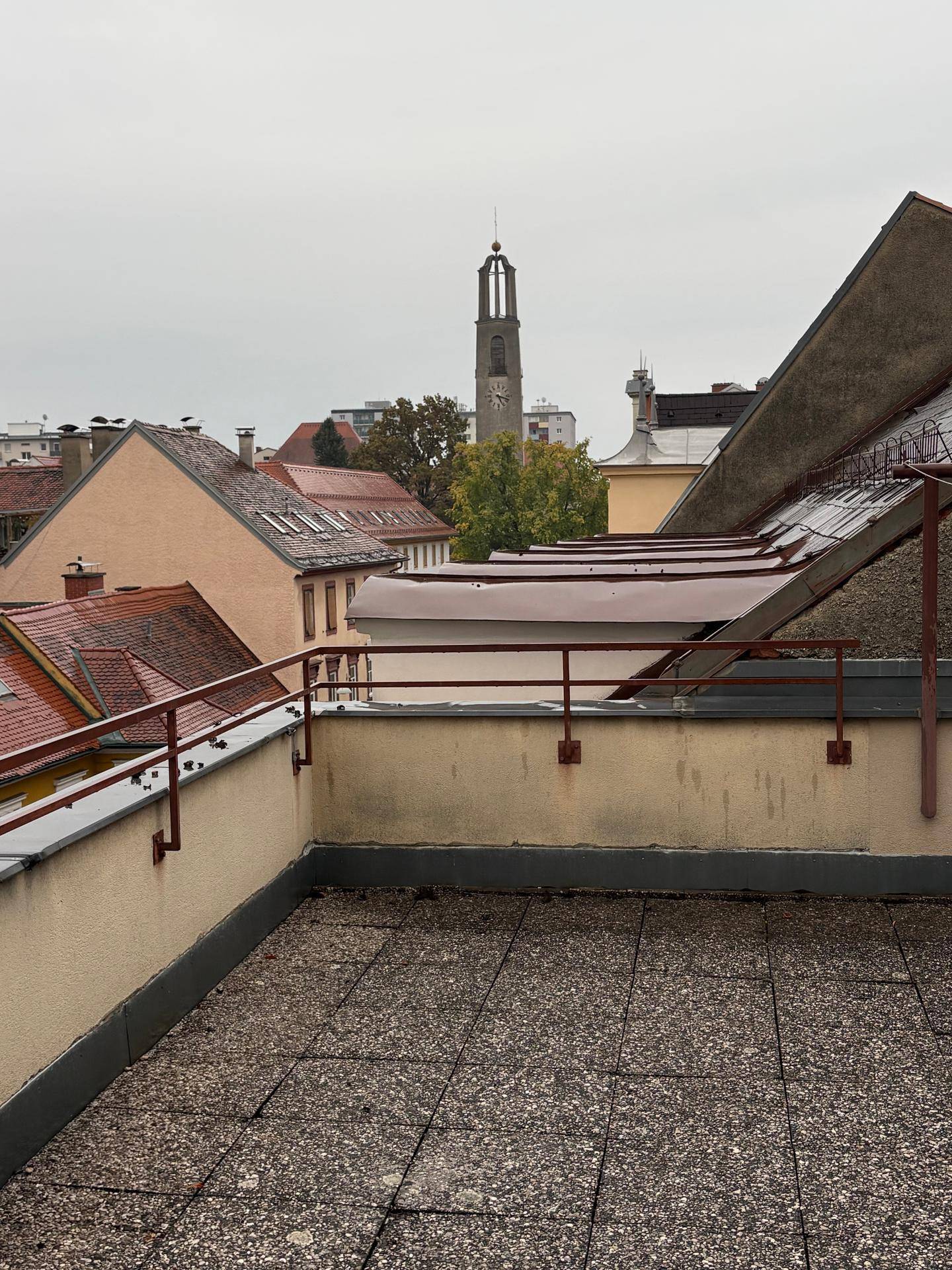 Geräumige Dachterrasse mit Fliesenboden und weitem Blick über die Stadt und einen Kirchturm.