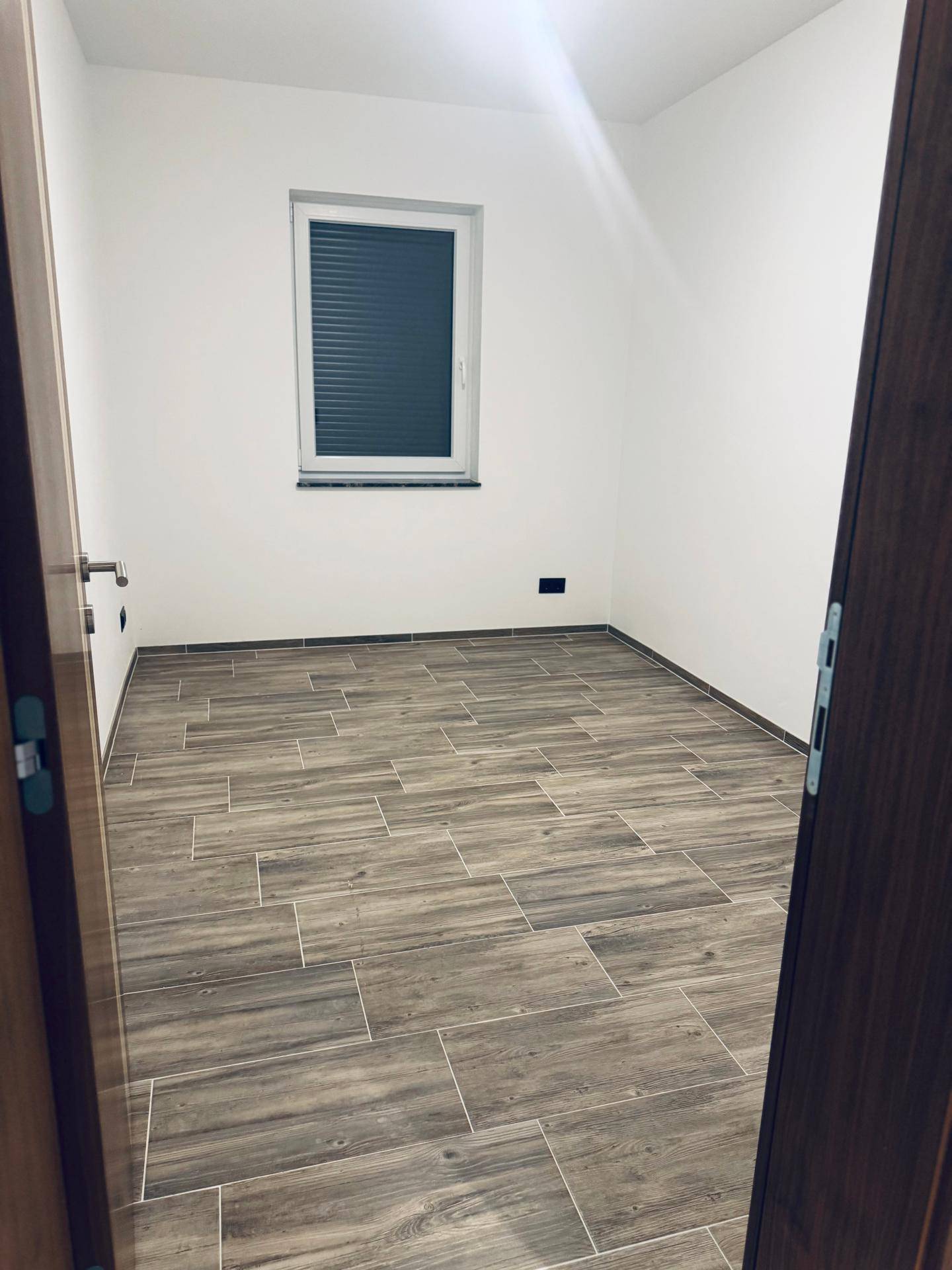 Helles Zimmer mit Holzoptik-Fliesenboden, weißen Wänden und einem Fenster mit geschlossenen Jalousien.