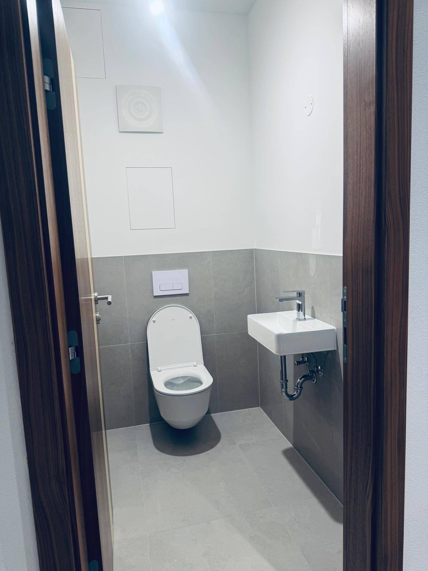 Modernes Gäste-WC mit wandhängender Toilette, kleinem Waschbecken und grauen Fliesen.