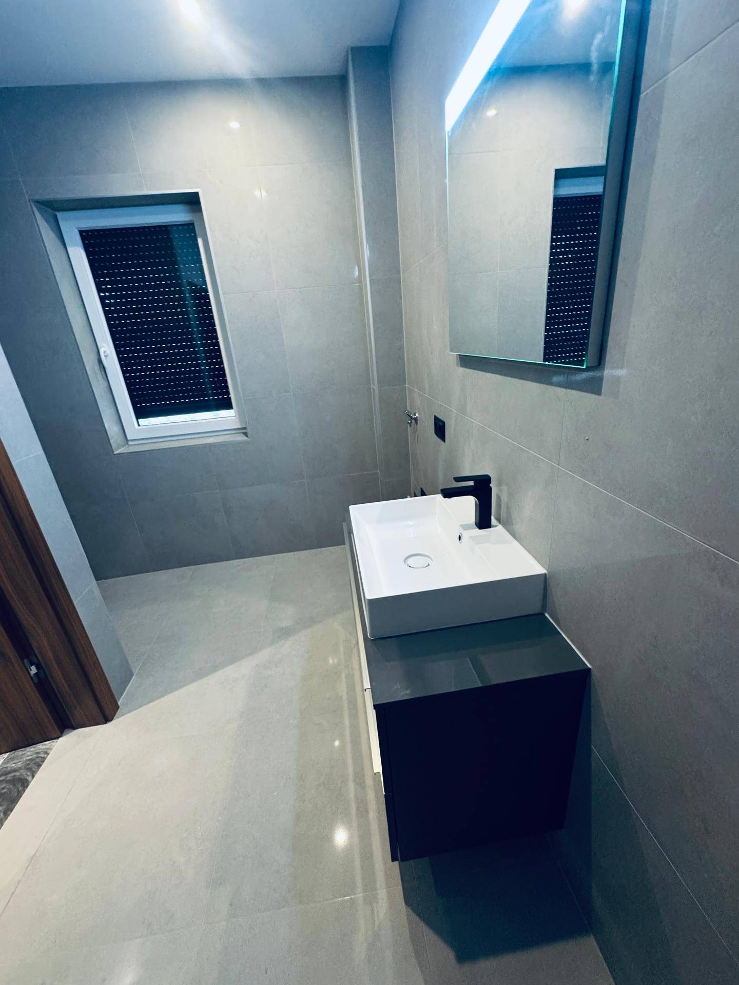 Helles, modernes Badezimmer mit Waschtisch, beleuchtetem Spiegel und Fenster mit Rollladen.