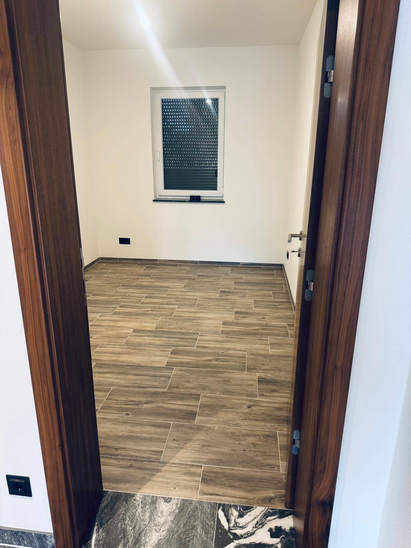 Geräumiges Zimmer mit ansprechendem Holzoptik-Fliesenboden und Fenster, ideal als Schlafzimmer.