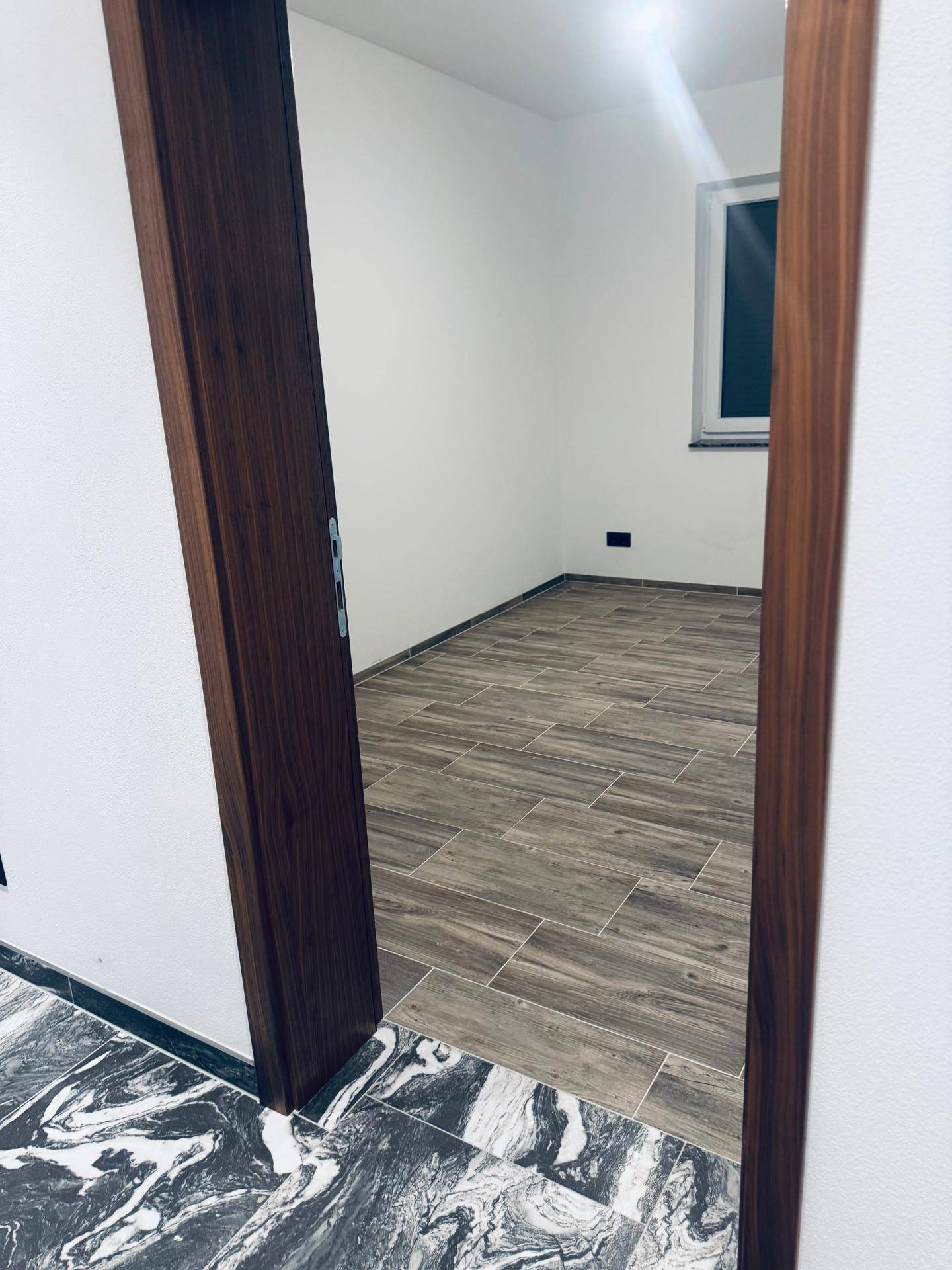 Blick vom Flur in ein helles Zimmer mit Holzoptik-Fliesenboden und einem Fenster.