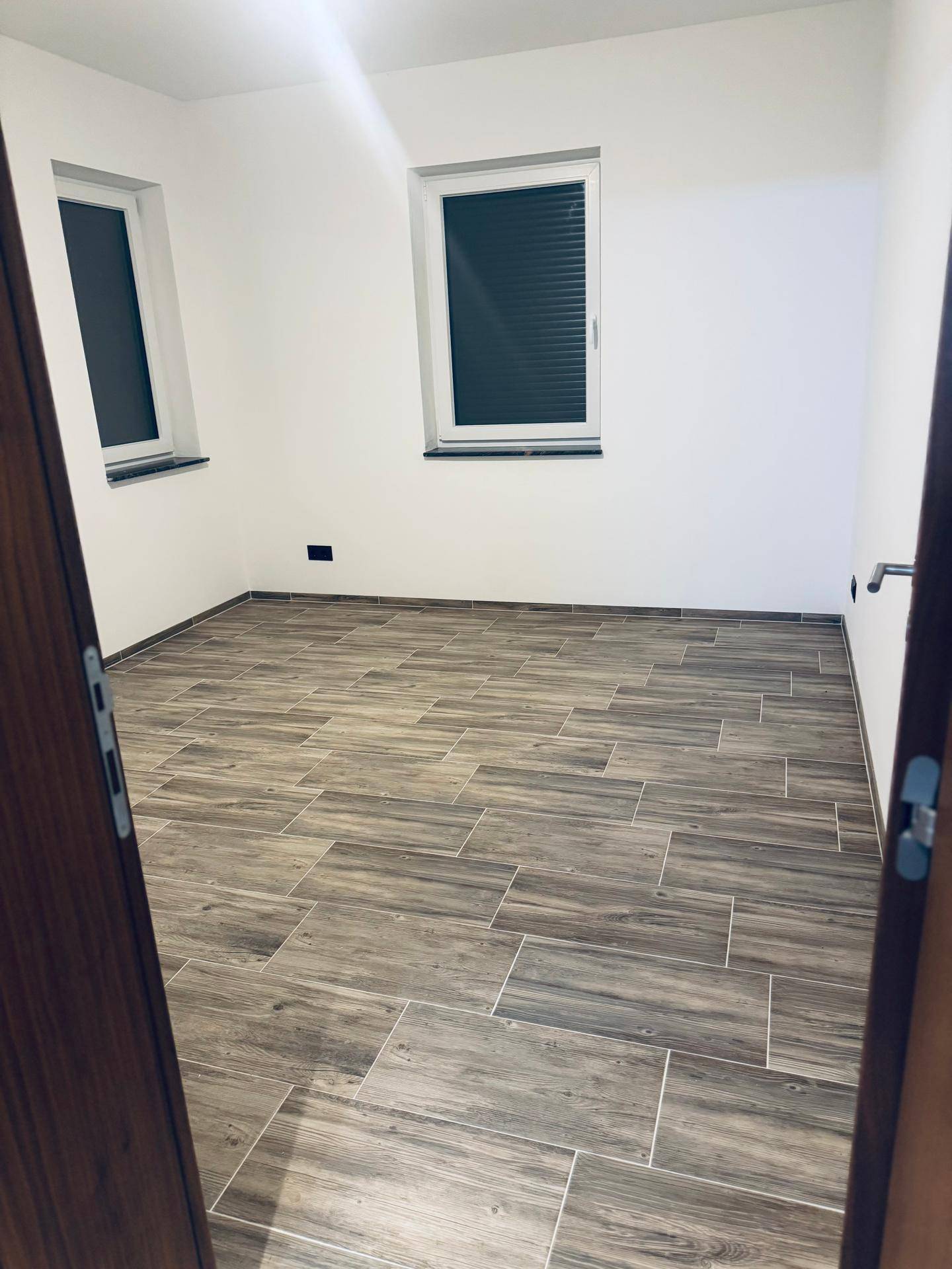 Leeres Zimmer mit Holzoptik-Fliesenboden, weißen Wänden und zwei Fenstern mit Jalousien.