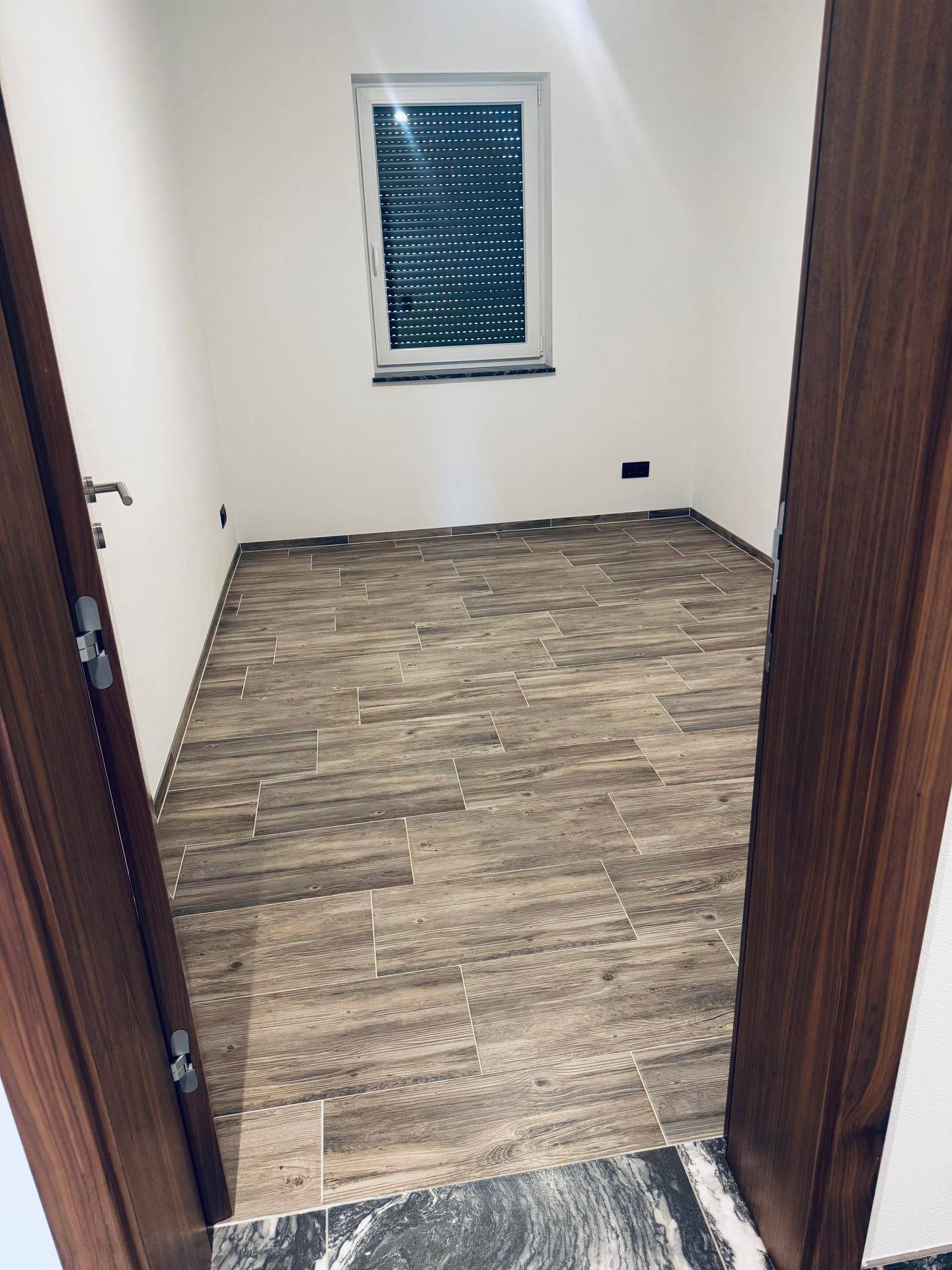 Helles Schlafzimmer mit Holzoptik-Fliesenboden und Fenster mit Rollladen, bereit zur Einrichtung.