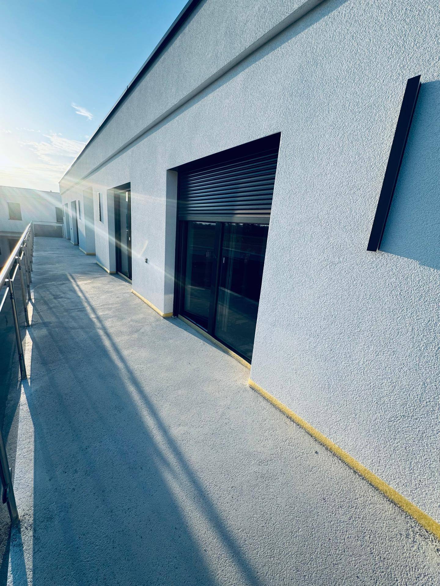 Moderne Gebäudefassade mit langem Balkon und schwarzen Rollläden unter strahlendem Himmel.