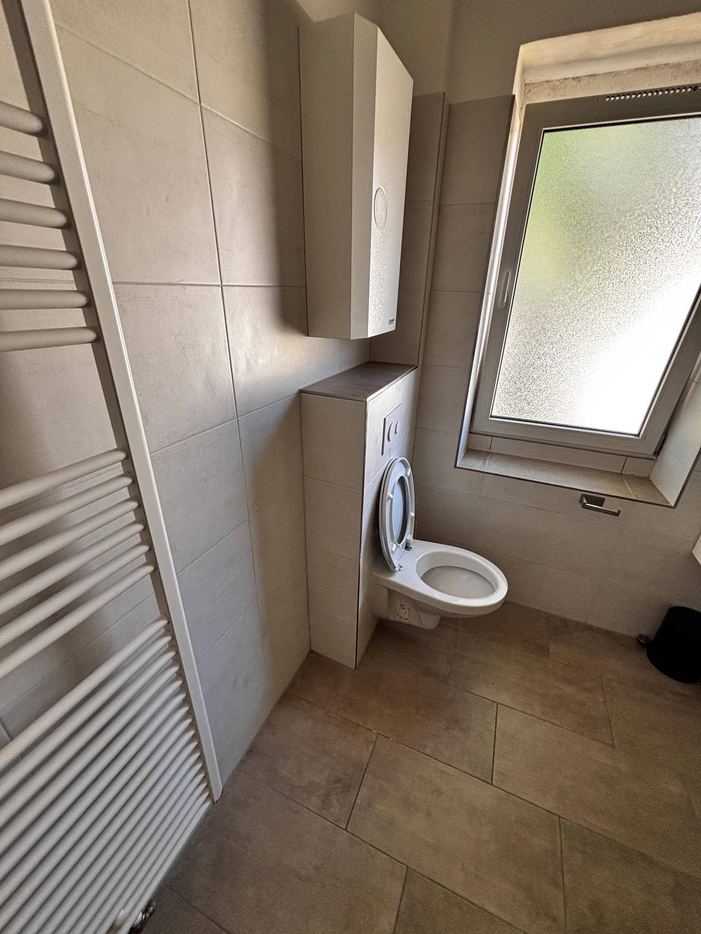 Badezimmer mit wandhängender Toilette, Handtuchheizkörper und Fenster.