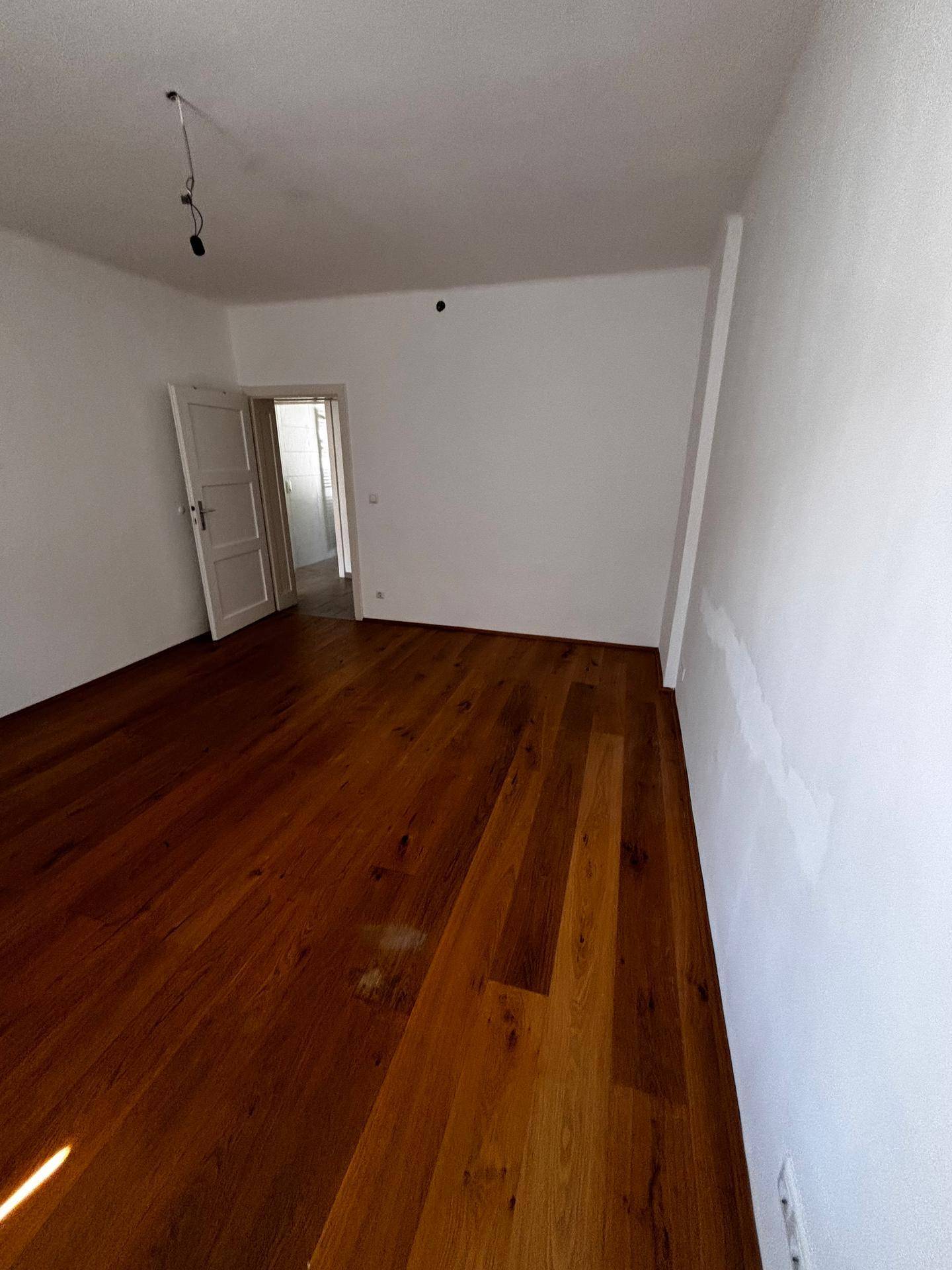 Leeres Zimmer mit warmem Holzboden und Zugang zu einem angrenzenden Bereich.