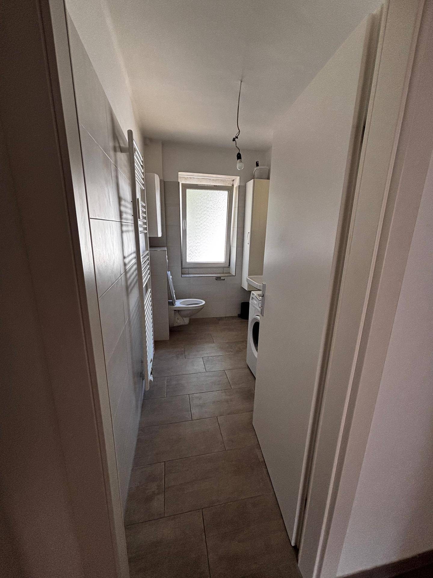 Badezimmer mit Fenster, Toilette und Waschmaschine, vom Flur aus sichtbar.