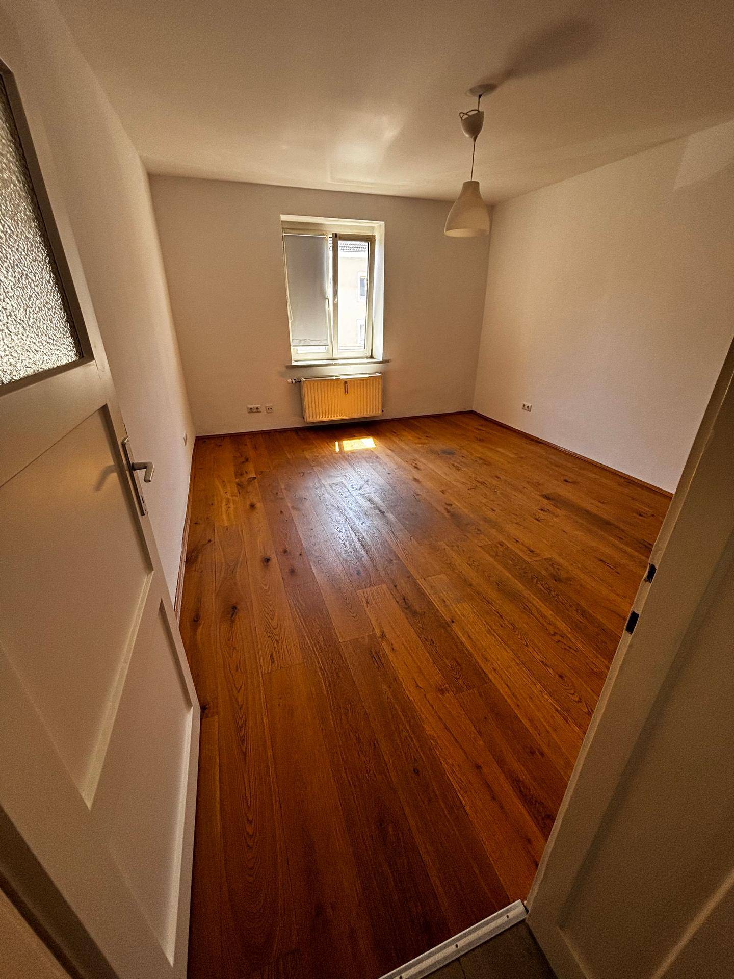 Helles, leeres Zimmer mit Holzboden, Fenster und Heizkörper, bereit zur Einrichtung.