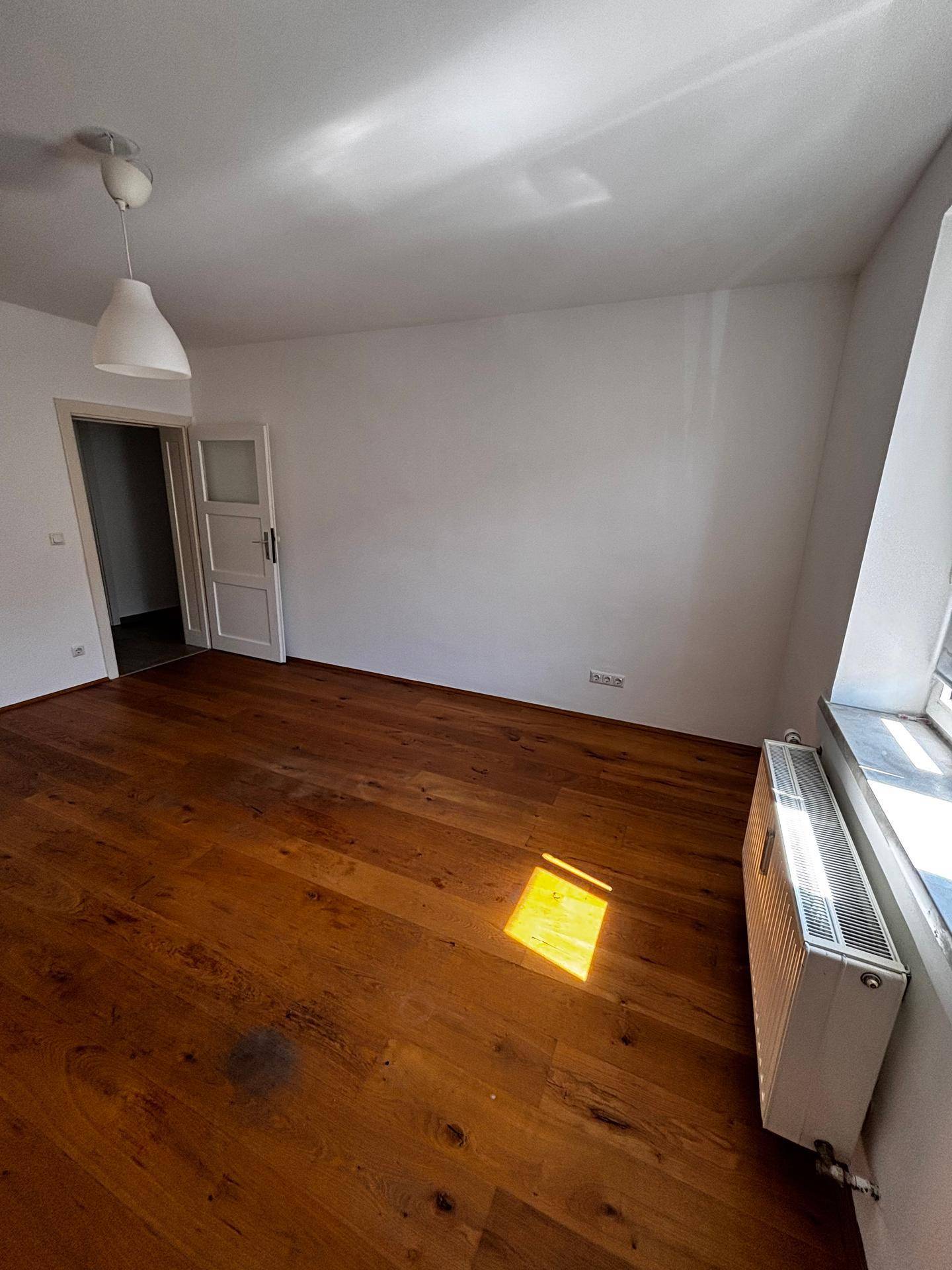 Sonniges, leeres Zimmer mit Holzboden, Fenster und Heizkörper, ideal für individuelle Gestaltung.