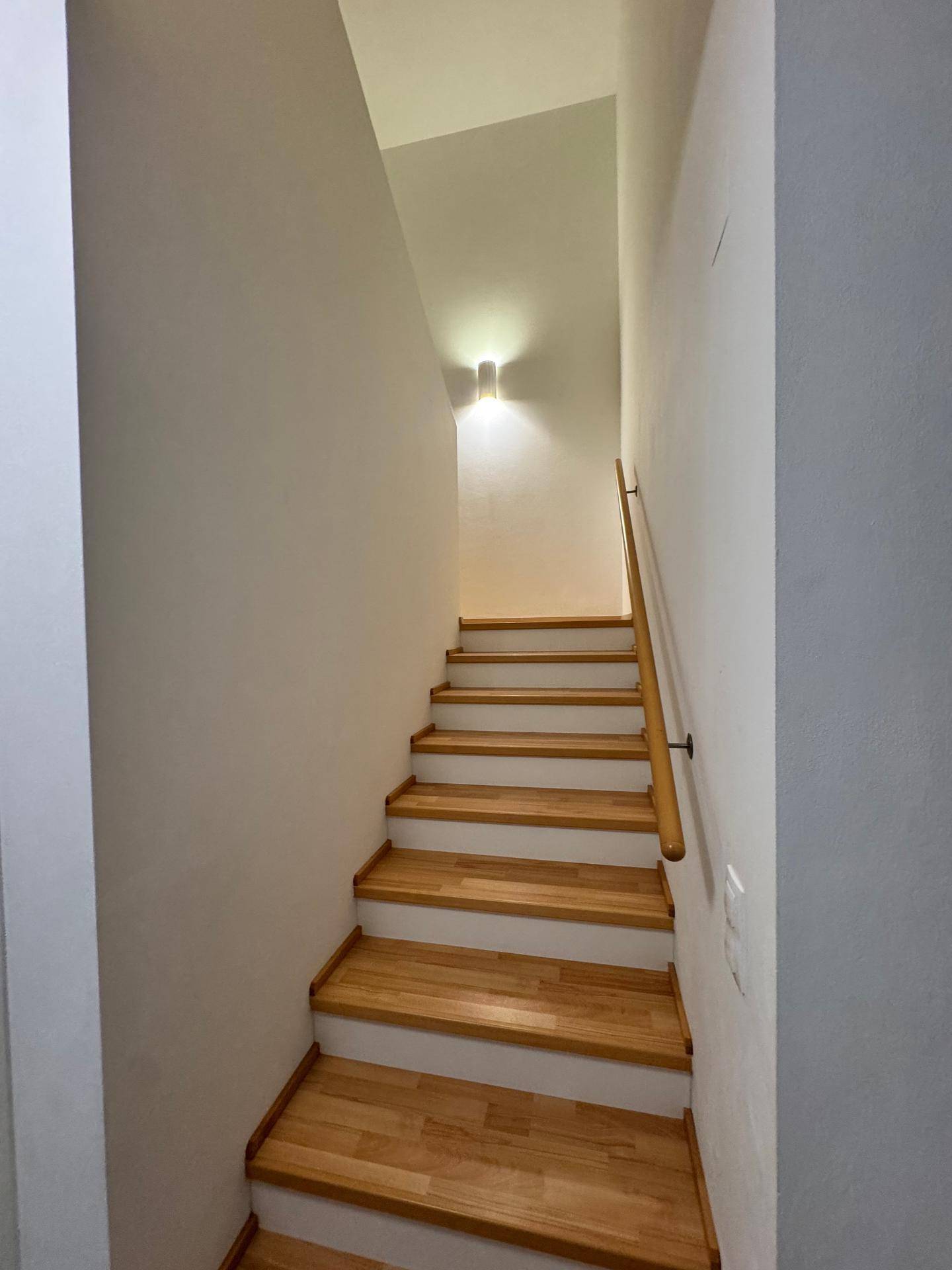 Treppenaufgang mit Holzstufen und hellem Geländer, beleuchtet von einer Wandleuchte.