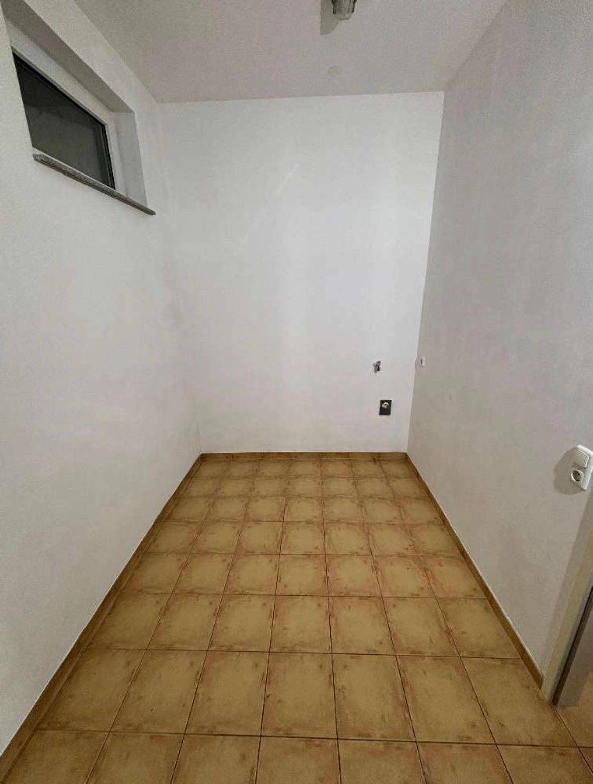 Kleiner Raum mit Fliesenboden und Fenster, vorbereitet für Waschmaschinenanschluss oder als Abstellraum.