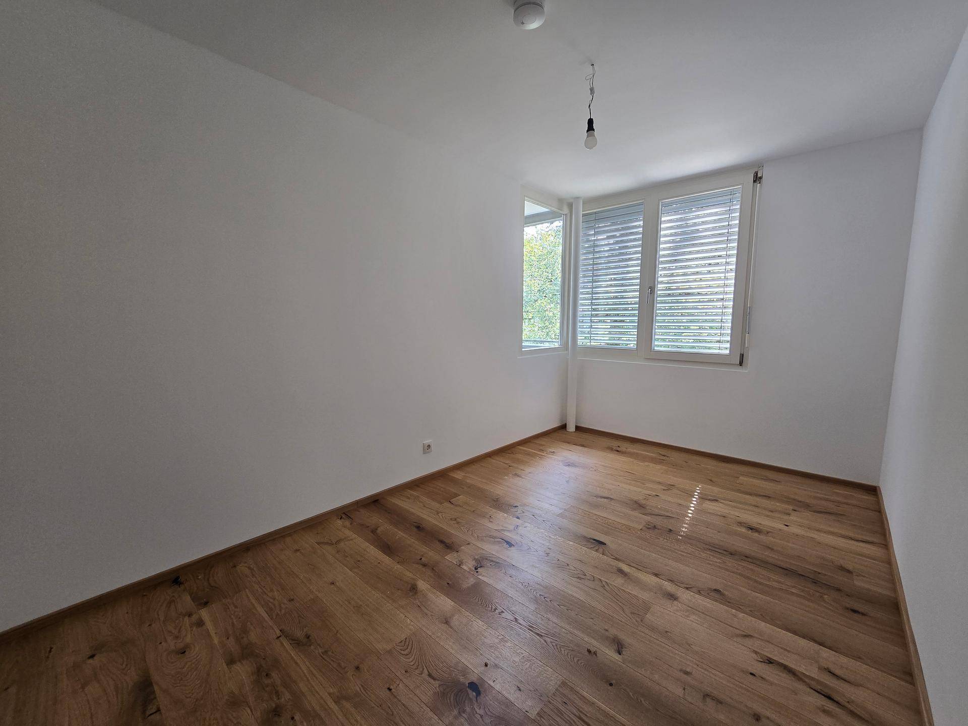 Helles Schlafzimmer mit großen Fenstern, Jalousien und hochwertigem Parkettboden.